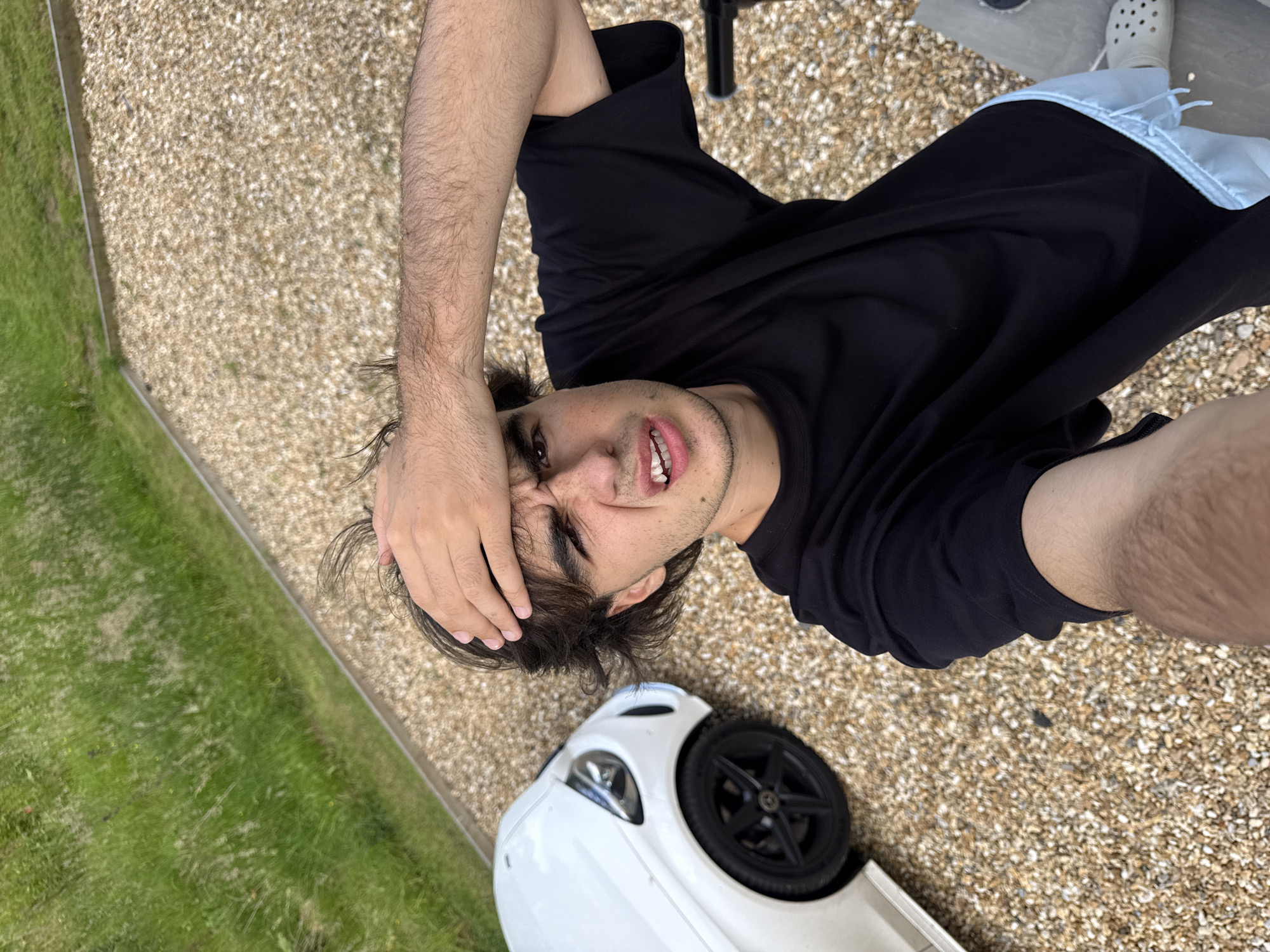 A young man is lying down on a gravel surface next to a white car. He is wearing a black t-shirt and light blue shorts. The man has his right arm raised and resting on his forehead, with his fingers spread. His eyes are closed and he appears to be grimacing or in pain. The car is a white sports car, and it appears to be upside down, with its wheels pointing upwards. The background consists of green grass and a gravel path. The lighting suggests it is daytime, and the weather appears to be clear. The man's expression conveys distress. There is no visible text in the image.