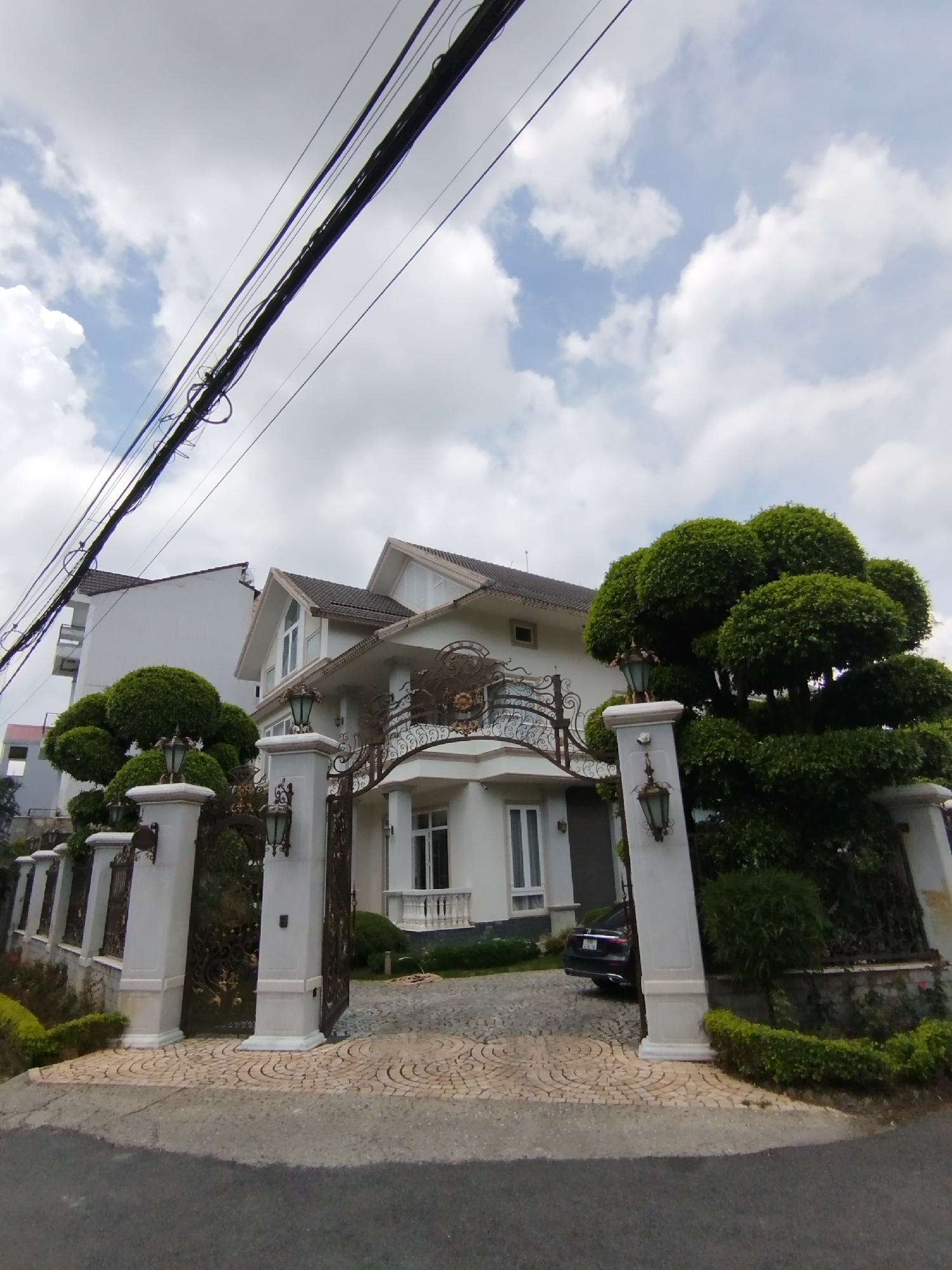 The image depicts a modern, multi-story house with a white facade and a dark tiled roof, located in Da Lat, Vietnam. The house is accessed through a grand entrance featuring ornate black metal gates and white stone pillars topped with decorative lamps. A dark-colored sedan is parked on the cobblestone driveway leading to the house. The property is surrounded by manicured greenery, including a large, spherical topiary tree to the right of the entrance. Overhead, power lines crisscross the sky, and a few scattered clouds are visible. The overall impression is one of wealth and meticulous landscaping.