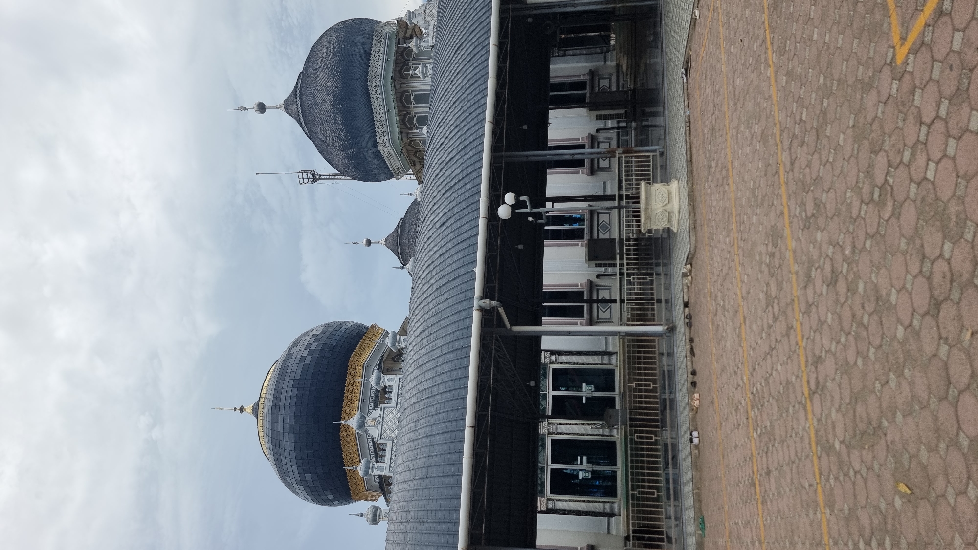 The image depicts the exterior of a mosque in Keudekruenggeukueh, Indonesia. The sky is overcast, with clouds dominating the upper portion of the frame. The mosque features prominent domes, one larger and black with gold accents, and another smaller, dark gray dome. A covered walkway with a dark gray, corrugated metal roof extends across the foreground, supported by white pillars. Glass doors and windows are visible behind the walkway railing. The ground in front of the walkway is paved with reddish-brown hexagonal interlocking bricks, marked with yellow lines. Several pairs of shoes are placed neatly on the pavement near the walkway. To the right of the walkway, a decorative white stone planter is positioned. The overall scene suggests a place of worship on a cloudy day.