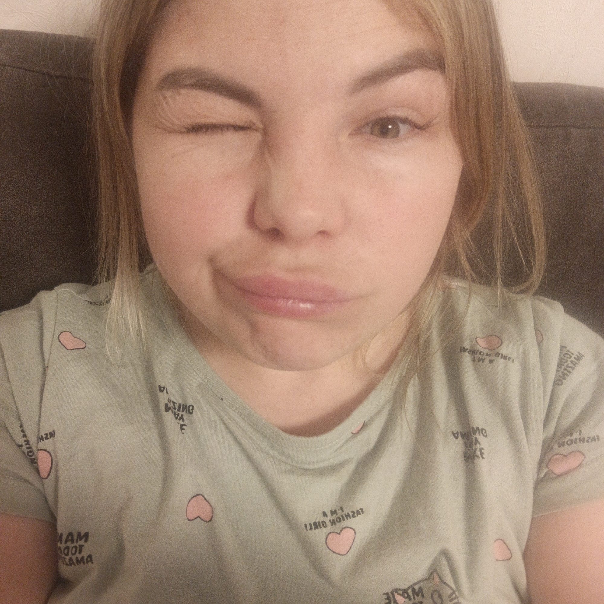A close-up, eye-level indoor shot features a light-skinned person with long, light brown hair, directly facing the camera. The subject is engaged in a playful expression, winking with their left eye and forming a pouting or "duck face" with their lips. Their open right eye is light brown or hazel, and the closed left eye exhibits prominent, long eyelashes.
The person is wearing a light green t-shirt patterned with small pink hearts and various lines of dark, reversed text. Visible fragments of text include "MAM AGOT AMA", "AMI JRID MOTHER", and "MOIMZAT", along with other scattered letters and numbers, suggesting the image may be a mirror selfie or has been horizontally flipped.
The background is simple, consisting of a dark, upholstered surface, possibly a couch or headboard, behind the subject's shoulders, and a lighter-colored wall or surface visible to the right. The lighting indicates an indoor setting, likely under artificial light. The overall mood conveyed is playful and lighthearted.