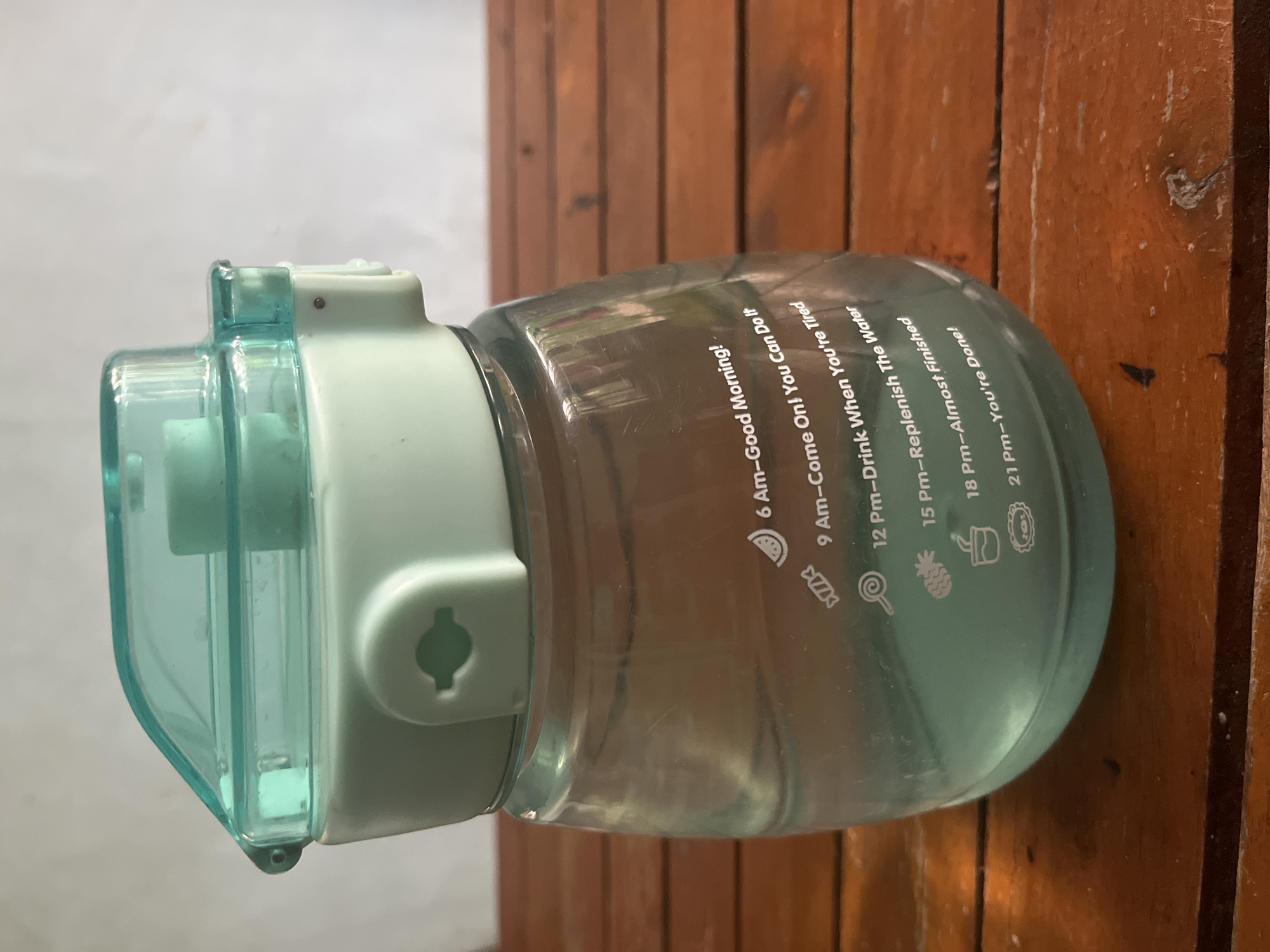 A mint green water bottle with a translucent teal lid sits on a wooden table. The bottle has time-based motivational markers printed on its side, including "6 Am-Good Morning!", "9 Am-Come On! You Can Do It", "12 Pm-Drink When You're Tired", "15 Pm-Replenish The Water", "18 Pm-Almost Finished", and "21 Pm-You're Done!". The water level is visible, reaching just above the "15 Pm" marker. The background is a plain, light-colored wall. The image is taken from a slightly elevated angle, with the wooden table surface occupying the foreground and lower portion of the frame. This scene does not contain any elements that would indicate a specific location in Meliau Hulu, Indonesia.