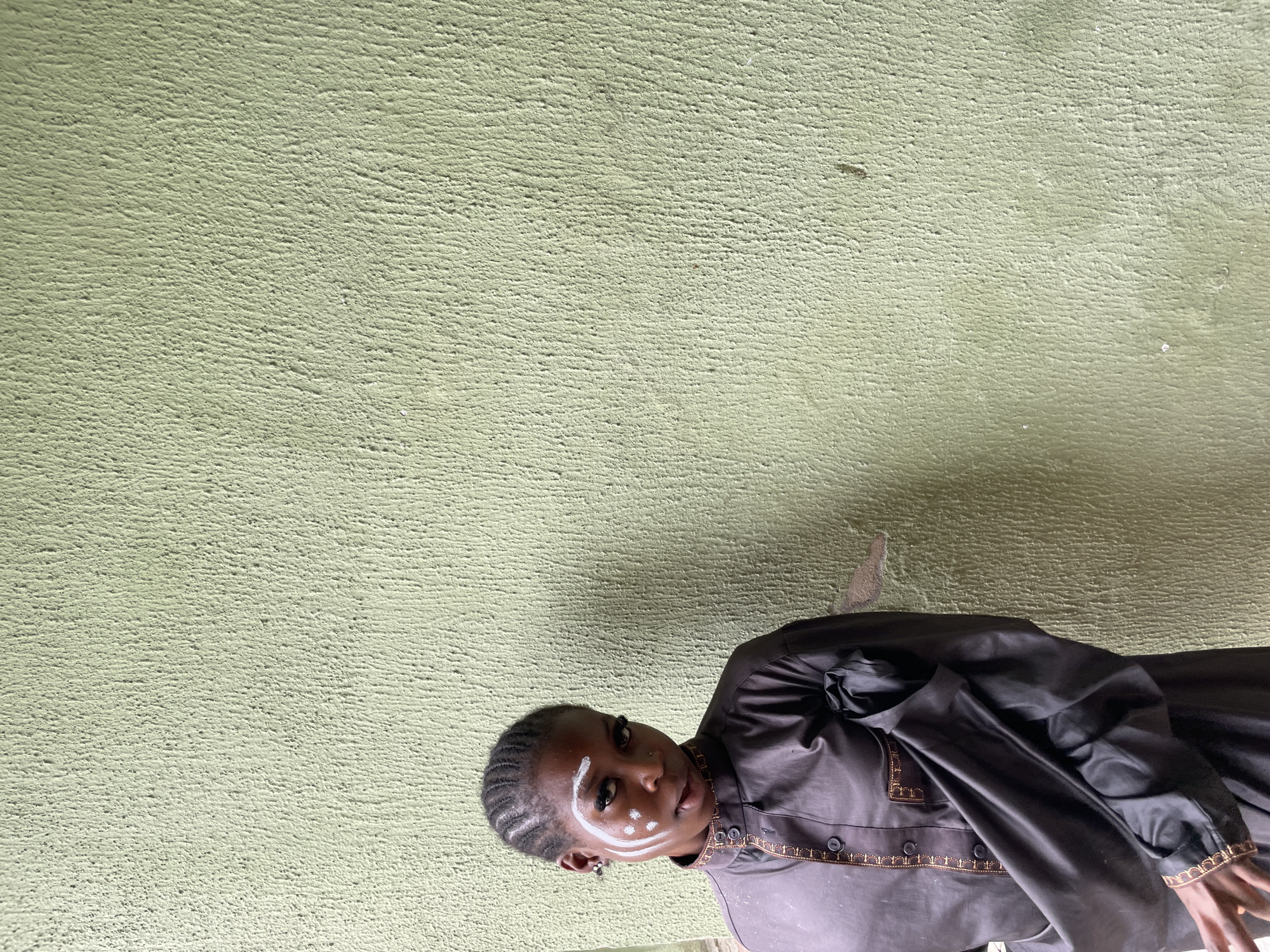 A young woman with braided hair and white face paint stands against a textured, pale green wall in Ado, Nigeria. She wears a dark grey garment with ornate gold trim along the collar and placket, and the sleeves are rolled up. The face paint consists of white markings around her eyes, including dramatic arcs above her eyebrows and dots on her cheeks. She is looking directly at the camera with a neutral expression. The wall behind her has a rough, stucco-like finish.