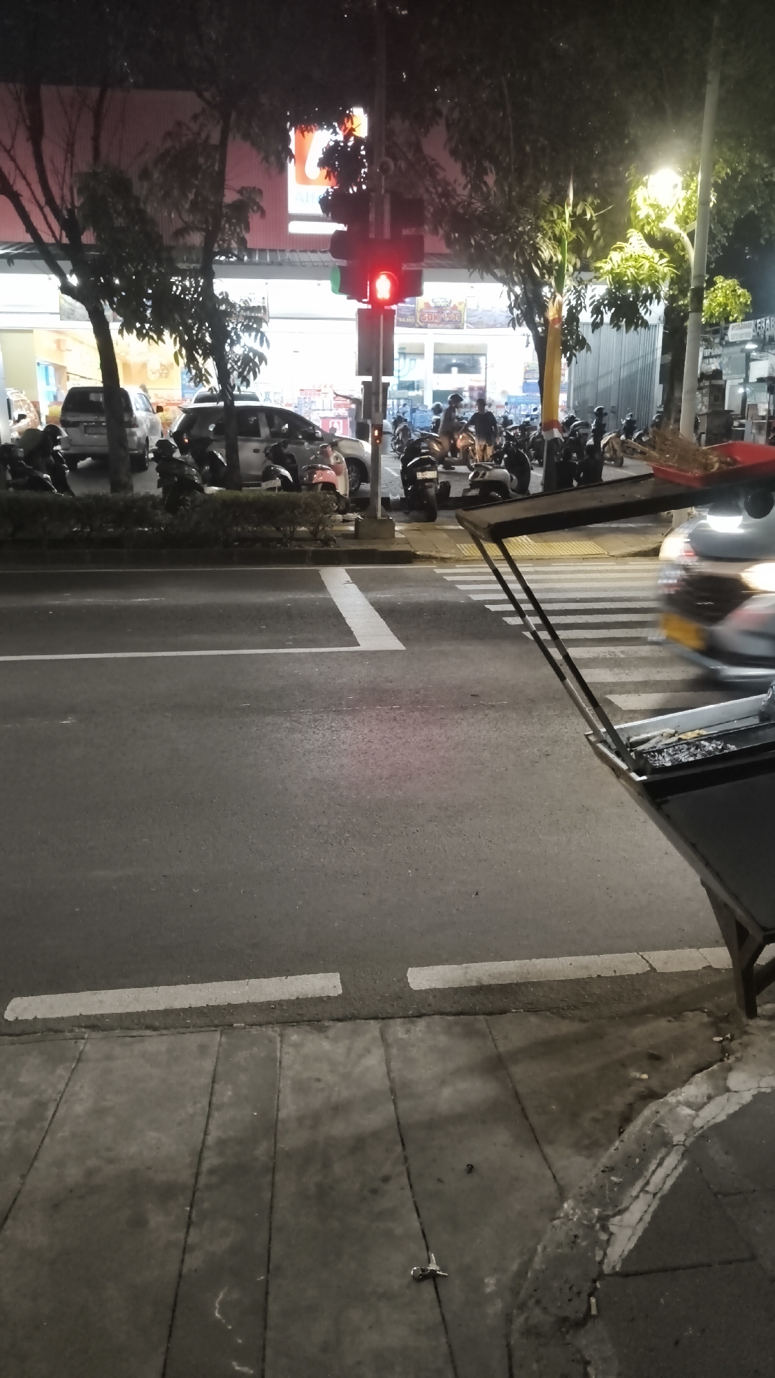 The image depicts a street scene at night in South Jakarta, Indonesia, likely in an urban commercial area. A traffic light displays a red signal. Numerous motorcycles are parked and several individuals are on or near them, suggesting a busy area. A food cart is visible in the foreground, equipped with a grill and food items. A car is in motion, with its headlights illuminated. The street is paved, with pedestrian walkways and crosswalks clearly marked. Trees line the street, and a building with illuminated signage is visible in the background.