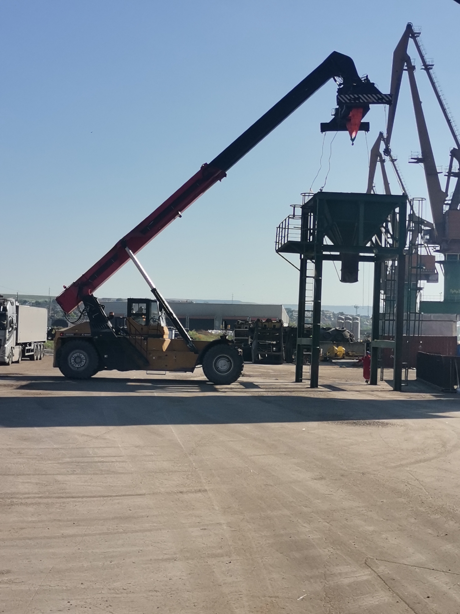 The image depicts a port scene in Ezerovo, Bulgaria, under a clear blue sky. A large, yellow and black telescopic handler is positioned in the foreground, its boom extended and poised above a metal hopper. The handler appears to be in the process of lifting a container or load. Adjacent to the hopper are several large, stationary dockside cranes, some painted in orange and green. A partially visible white truck is parked on the left. There is a building in the background, with an area of vegetation. A person wearing red stands near the hopper, presumably observing or assisting with the lifting operation. The ground is a concrete or paved surface.