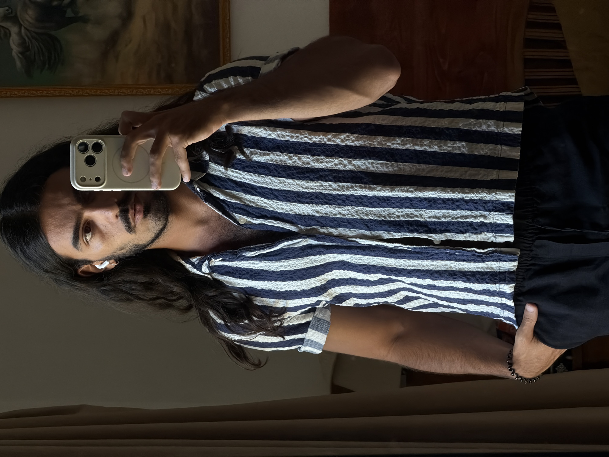A man is taking a selfie in a room. He is wearing a striped blue and white shirt and black shorts. He has long dark hair and a beard and is wearing an earpiece in his left ear. He is holding a smartphone with a tan case. In the background, there is a painting on the wall and a reflection in a mirror. The location is likely indoors, possibly in a residence or hotel in North Kuta, Indonesia.