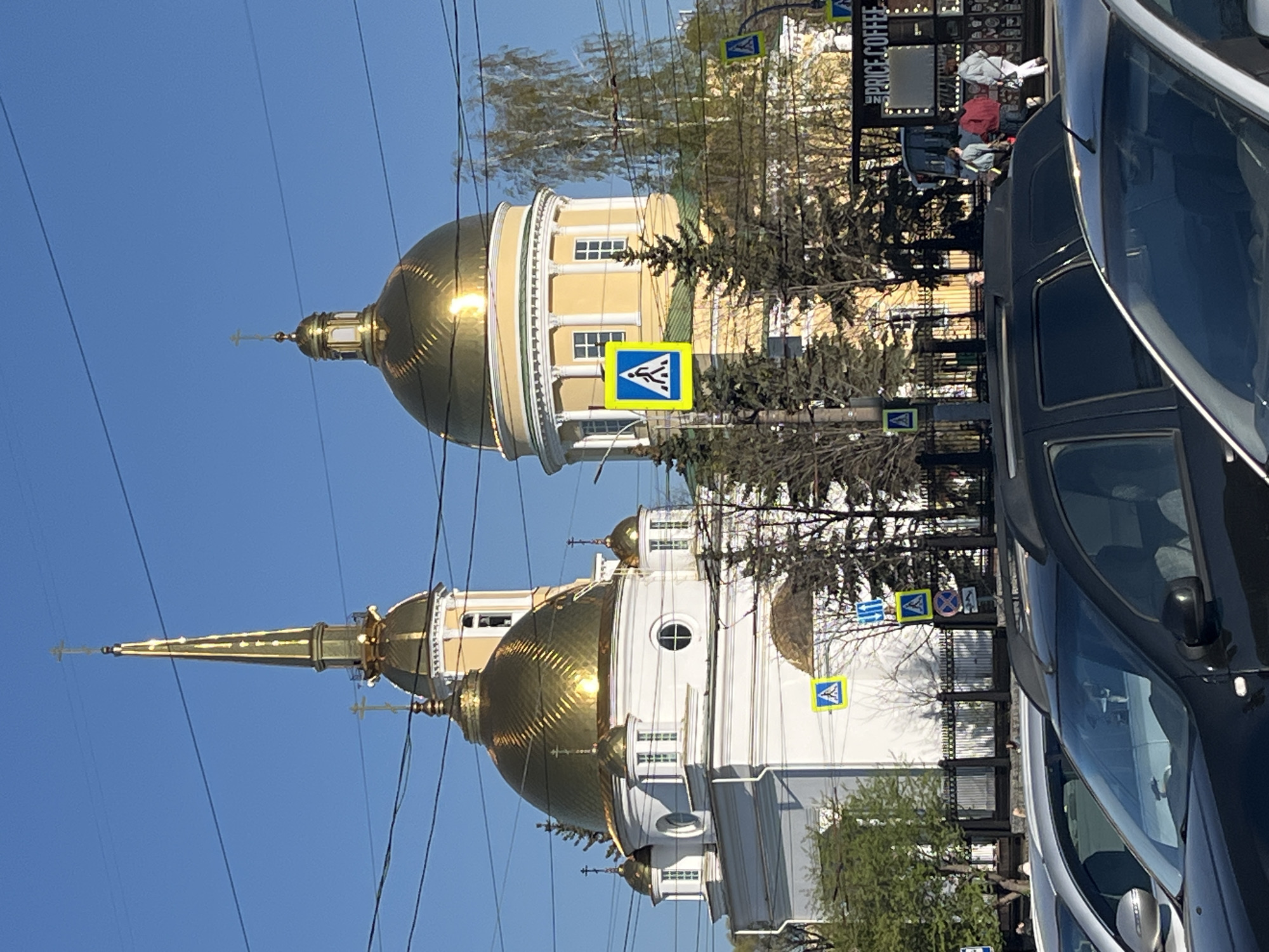 The image captures the Alexander Nevsky Cathedral, located in Izhevsk, Russia. The photograph showcases the cathedral's golden domes against a clear blue sky. The cathedral's architecture, featuring white walls and ornate details, is partially visible. Street signs are positioned in the foreground, along with the partial view of parked cars. The photograph is taken from street level. Pedestrians and possibly a coffee stand are visible along the sidewalk adjacent to the street. Utility wires crisscross the sky above the cathedral.