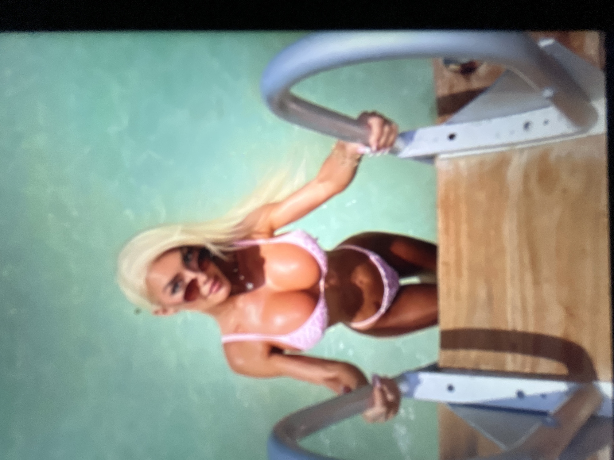 A woman with blonde hair and sunglasses is climbing out of turquoise water using a metal and wood ladder. She is wearing a pink bikini. The background is a blurred turquoise surface. The scene is not located in Sofia, Bulgaria.
