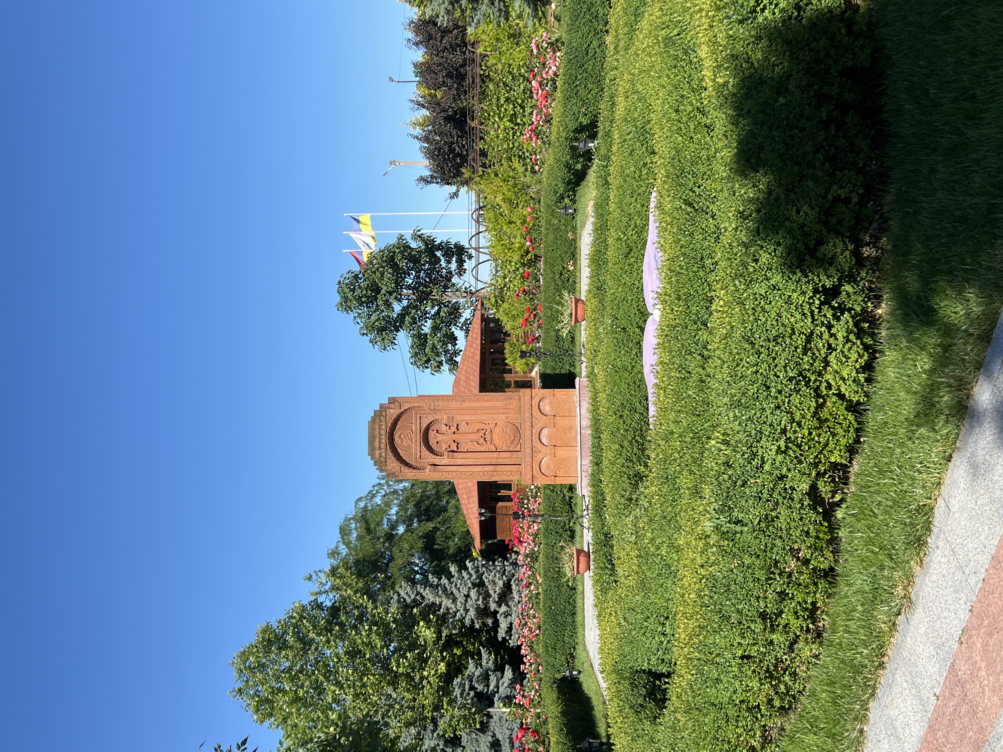The image shows a beautifully landscaped garden with a prominent stone monument in the center. The monument is reddish-brown and intricately carved with religious motifs. Surrounding the monument is a manicured circular hedge, with a pathway leading through it. Beyond the hedge, a vibrant bed of red and pink roses blooms, with a backdrop of lush green trees and a small building with a red roof. Several flags are visible in the distance, fluttering against a clear blue sky. The scene appears to be a tranquil and well-maintained outdoor space, likely a park or a religious site. The presence of flags, specifically the Ukrainian flag, suggests the location is in Ukraine, and the mention of Odesa, Ukraine, in the prompt indicates this specific city as the setting. The image is taken from a slightly elevated perspective, looking across the garden.