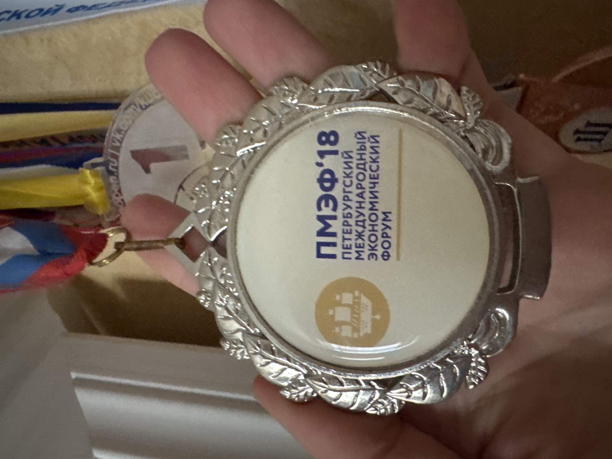 The image shows a close-up of a hand holding a medal. The medal is circular with a silver-toned decorative border featuring leaf motifs. The central part of the medal is white with text and a graphic. The text is in Russian and appears to read "ПМЭФ'18" at the top, followed by "ПЕТЕРБУРГСКИЙ" and "МЕЖДУНАРОДНЫЙ" on subsequent lines, and then "ЭКОНОМИЧЕСКИЙ" and "ФОРУМ" at the bottom. There is a yellow circular emblem to the left of the text, containing a silhouette of a sailing ship. Above the medal, a ribbon in red, white, and blue is visible, suggesting it is a commemorative award. The background is out of focus but suggests an indoor environment with some light-colored surfaces. The medal appears to be from the "Petersburg International Economic Forum '18". The user specified the location as Sinyavino, Russia, but there is no visual information in the image to confirm or deny this location.