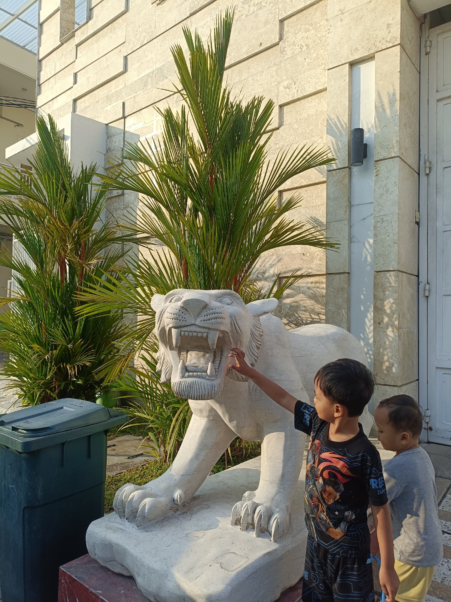 In Kediri City, Indonesia, two young boys are standing next to a large, white stone sculpture of a lion. The sculpture is positioned in front of a modern building with light-colored stone cladding and large glass windows. Lush green palm trees flank the sculpture. One boy, wearing a black t-shirt with a graphic print and patterned shorts, is reaching out to touch the lion's mouth. The other boy, wearing a grey t-shirt and yellow shorts, is standing beside him, looking at the sculpture. A dark green trash can is visible to the left of the scene. The lighting suggests it is daytime, possibly late afternoon, with shadows cast by the palm leaves on the building.