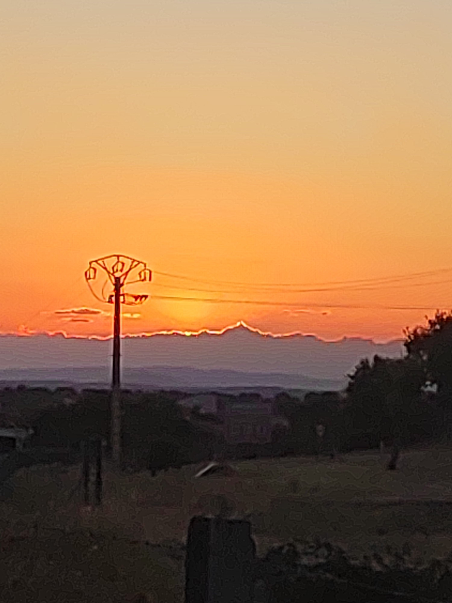 A low-angle shot captures a sunset over the landscape near Sobradillo, Spain. The sky transitions from a pale yellow at the top to a vibrant orange near the horizon. A solitary electrical pylon with wires stretching across the frame stands silhouetted against the sky. Below the horizon, a range of mountains is visible, their peaks softened by atmospheric haze. The foreground is dominated by dark, out-of-focus foliage and what appears to be a fence. The overall impression is one of a serene, rural evening.