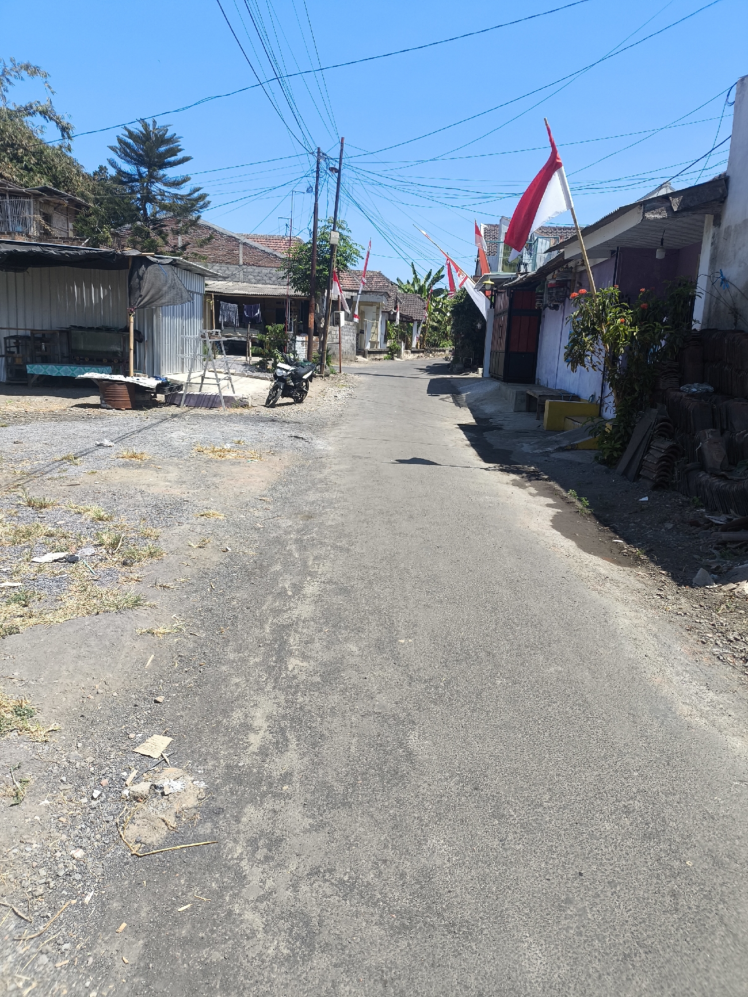 In Kebonsari, Indonesia, a sun-drenched, narrow asphalt road winds through a residential area. The sky is a clear, vibrant blue, with a tangle of overhead power lines crisscrossing the frame. Along the sides of the road, houses with red-tiled roofs and white or grey walls are visible. Many of these homes are adorned with Indonesian flags, red and white, flying from poles, indicating a national celebration. Some structures appear to be makeshift stalls with corrugated metal roofs. In front of one house, a motorcycle is parked on the gravelly shoulder. The environment is dusty, with dry grass and scattered debris along the road's edge. The scene captures a quiet street life, possibly during a holiday or event, with a clear focus on the local architecture and the ubiquitous presence of Indonesian national symbols.