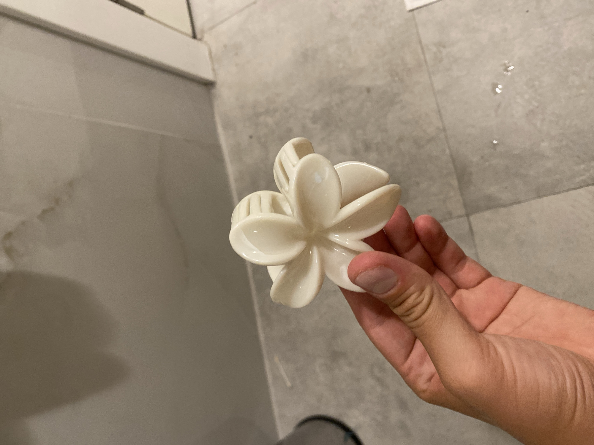 A hand holds a white, flower-shaped hair claw in front of a tiled floor and wall. The claw has five petals and a ribbed center. The hand is tanned with short fingernails. The background appears to be a bathroom or similar tiled environment with a wall and floor. There are some water droplets on the floor.