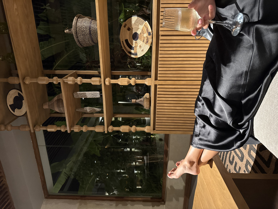 A rotated view captures a person seated, legs extending towards the bottom of the frame, barefoot. The individual wears a dark, silky garment. A partially filled wine glass is held in the upper right. Behind the person, an intricately designed wooden shelf displays woven baskets and decorative items. A large window showcases lush, dark greenery and a blurred building exterior at night. The interior lighting casts shadows, creating a warm, inviting atmosphere.