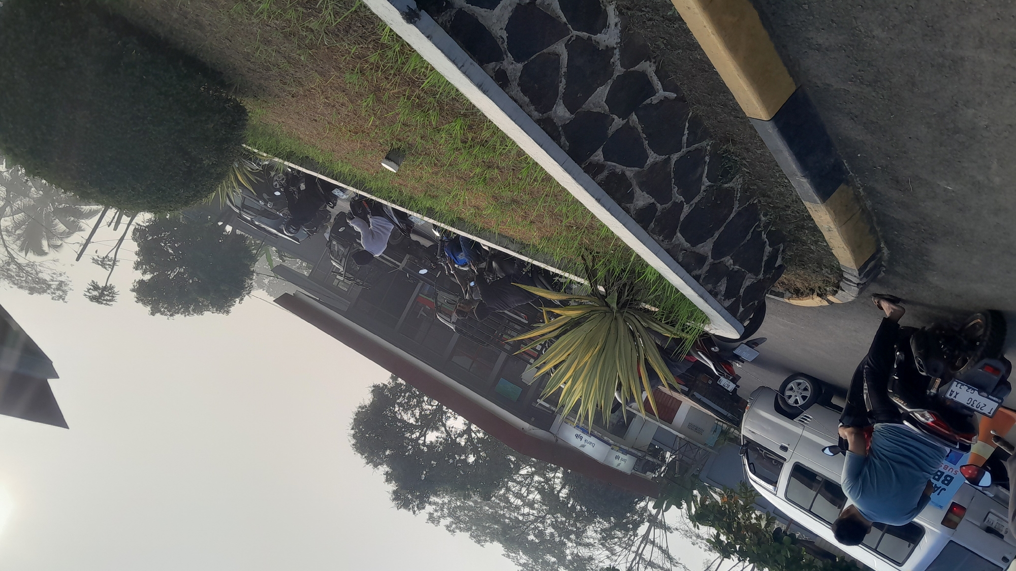 The image captures a street scene in front of a building, likely a bank or commercial establishment, in Subang, Indonesia. The foreground features a paved area with a decorative stone wall and a patch of grass. To the left, a man wearing a blue t-shirt and dark pants is seated on a black scooter, facing away from the camera. He is wearing flip-flops. A white van is parked behind him. Further back, several other scooters and a black car are parked. A building with a red roof and windows is visible in the background, with signage that appears to read "bank bjb". The atmosphere is hazy, suggesting either morning mist or pollution. Trees, including palm trees, are scattered throughout the scene, and the sky is overcast with the sun peeking through on the right side. The overall impression is one of a typical urban environment in Indonesia, possibly during the early morning or late afternoon due to the lighting and haze.