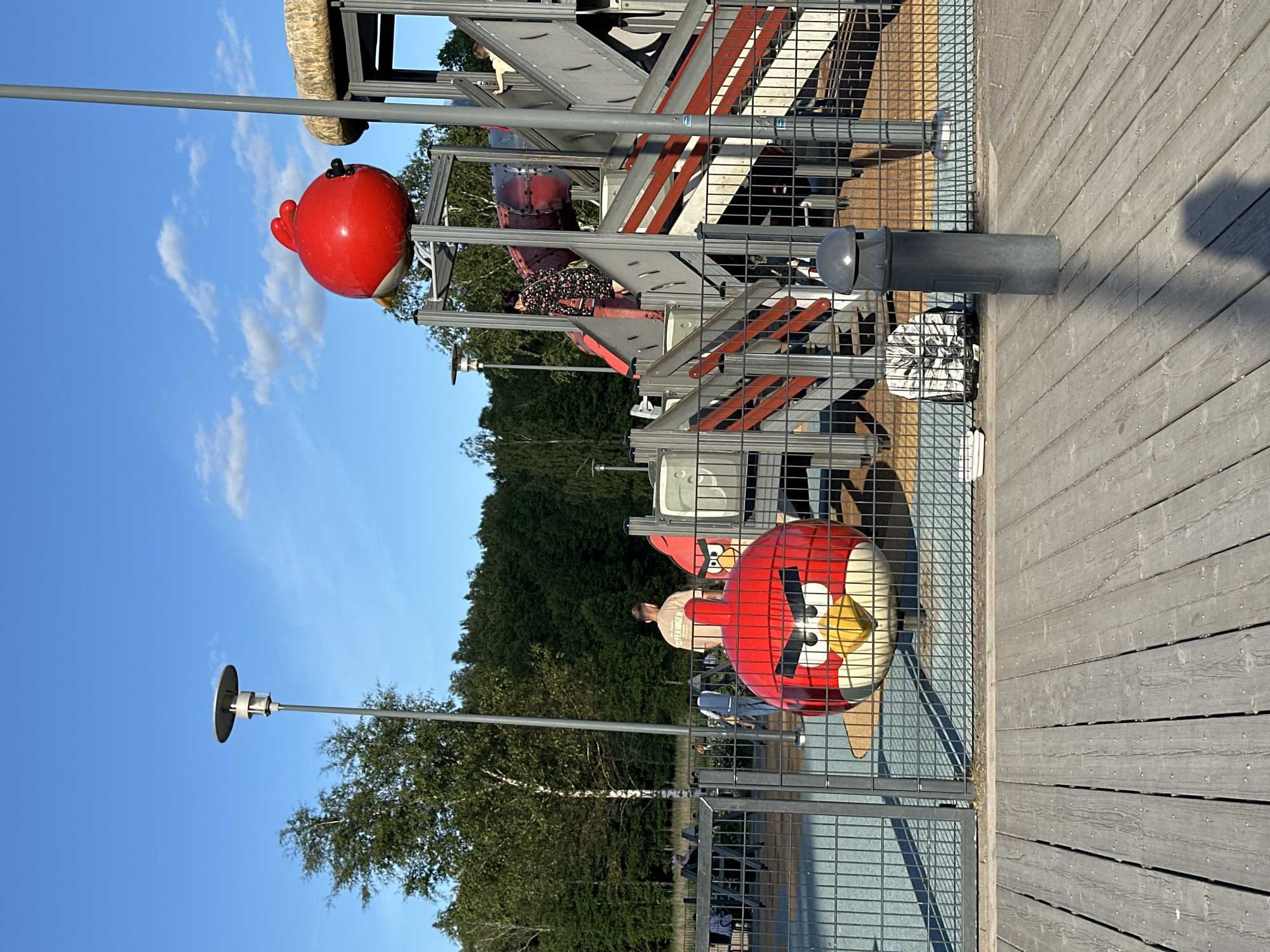 This is a medium shot, eye-level outdoor photograph taken on a sunny day with a clear blue sky and scattered clouds. The foreground features a weathered wooden deck. Beyond the deck is a black metal fence, behind which is a playground. Dominating the playground area are two large, red Angry Birds characters. One is positioned closer to the fence, facing forward with its signature angry expression. The other is perched higher up on a playground structure. The playground structure itself is made of grey metal with red accents and includes slides and stairs. Several people are visible in the background, some on the playground and others walking in the distance. Trees and foliage form a dense backdrop behind the playground. A tall lamppost stands to the left of the frame, and another grey pole, possibly a trash can or bollard, is located in the middle ground.