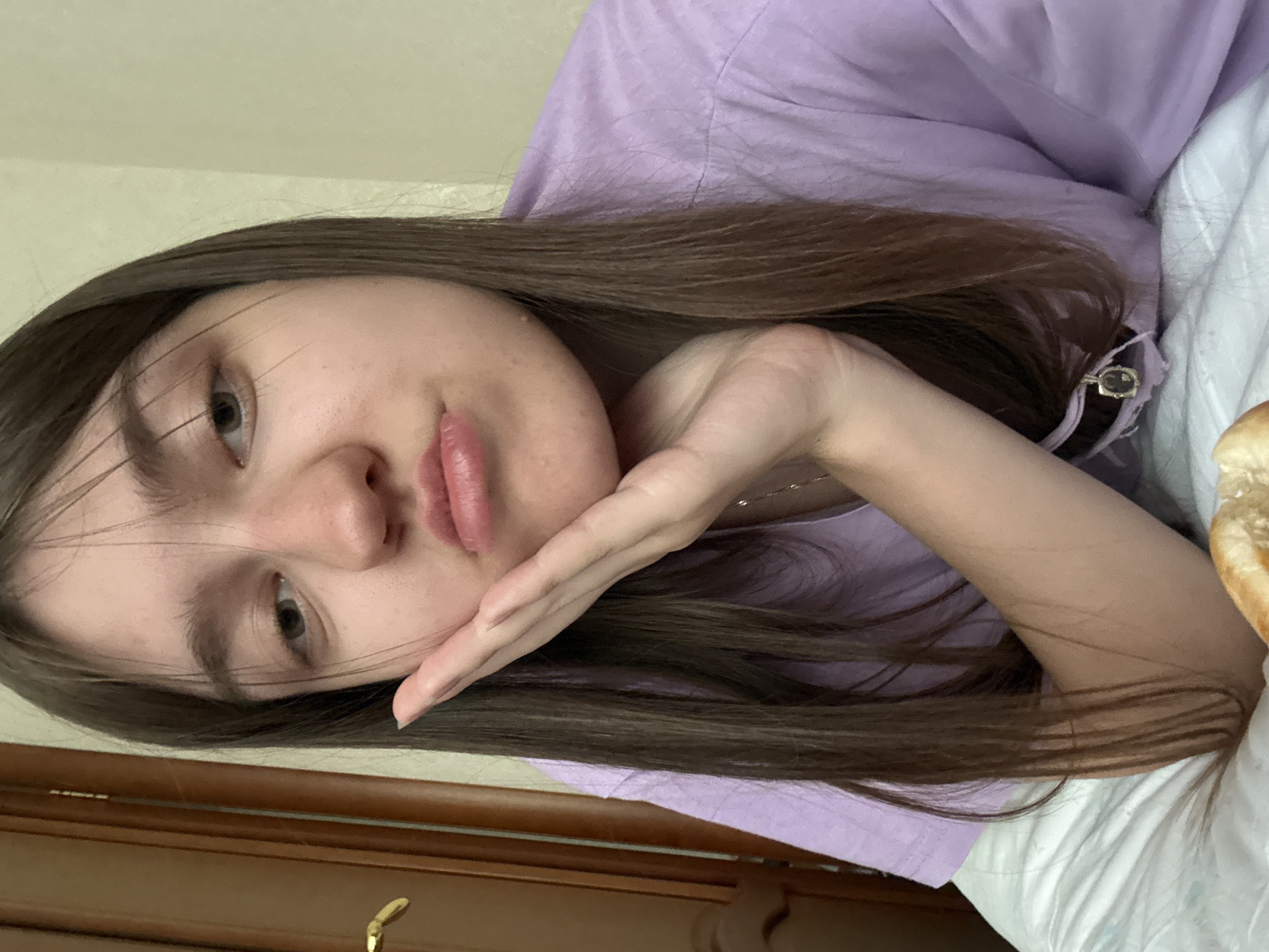 A young woman with long, dark brown hair is lying down, possibly on a bed, with a light purple t-shirt. She is resting her face on her hand, looking towards the camera. A bed or other soft surface is visible in the background, and a door or furniture item is visible at the bottom of the frame. The location is Unknown, Unknown.
