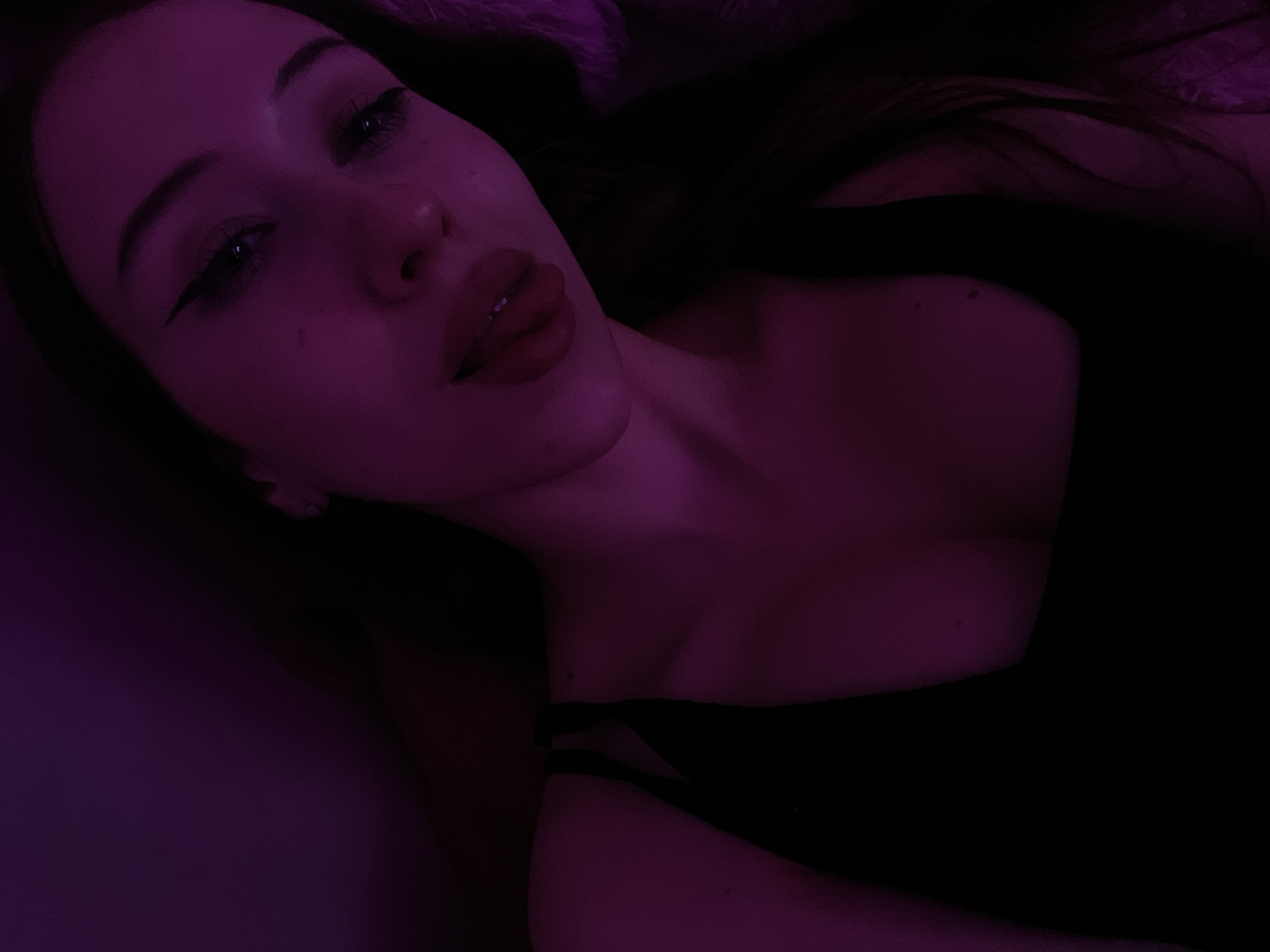 The image is a close-up, low-angle, selfie-style photograph of a young woman. She is positioned in the foreground, with her face angled towards the camera. The lighting is predominantly purple, casting a dim and atmospheric glow on the scene.
The woman has dark hair that falls around her shoulders and down her back. Her eyes are visible, with dark eyeliner accentuating them. Her lips are slightly parted, revealing her tongue, and appear to be wearing lipstick. She is wearing a black garment that is visible on her chest and shoulders.
The background is largely dark and out of focus, with hints of what might be a textured surface or fabric. The overall impression is intimate and moody, possibly taken in a bedroom or a dimly lit environment. There is no indication of the city of Votkinsk, Russia, within the image itself.