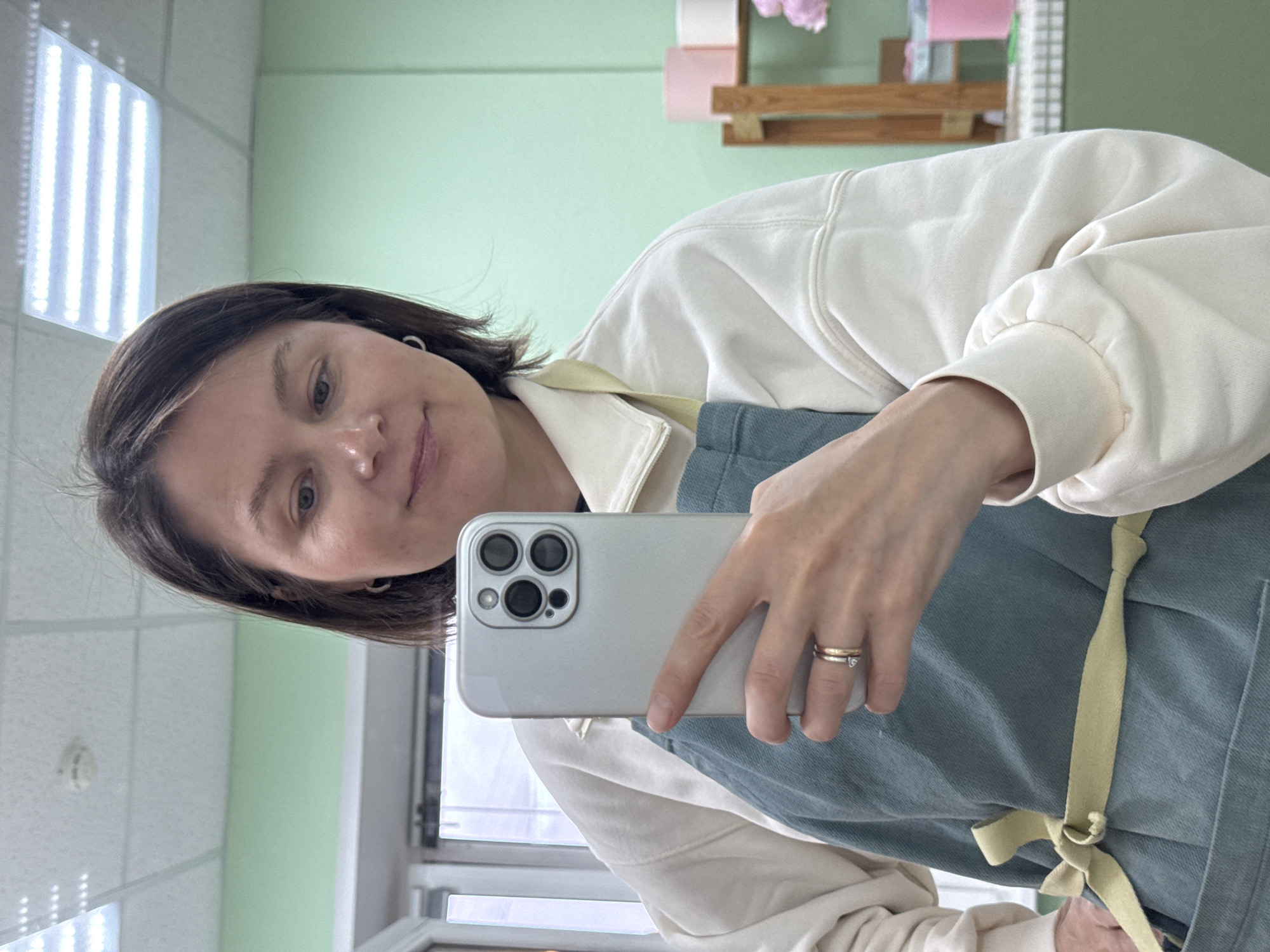 A woman is taking a selfie in what appears to be an indoor space. She is holding a silver smartphone in her right hand. The woman has short, dark hair and is wearing a light-colored sweater and a blue apron with a yellow tie. There are two rings visible on her finger. The background includes a pale green wall, a window frame, and what seems to be a shelf with objects on it. The room has fluorescent lighting. There is no indication in the picture that the location is in Izhevsk, Russia.