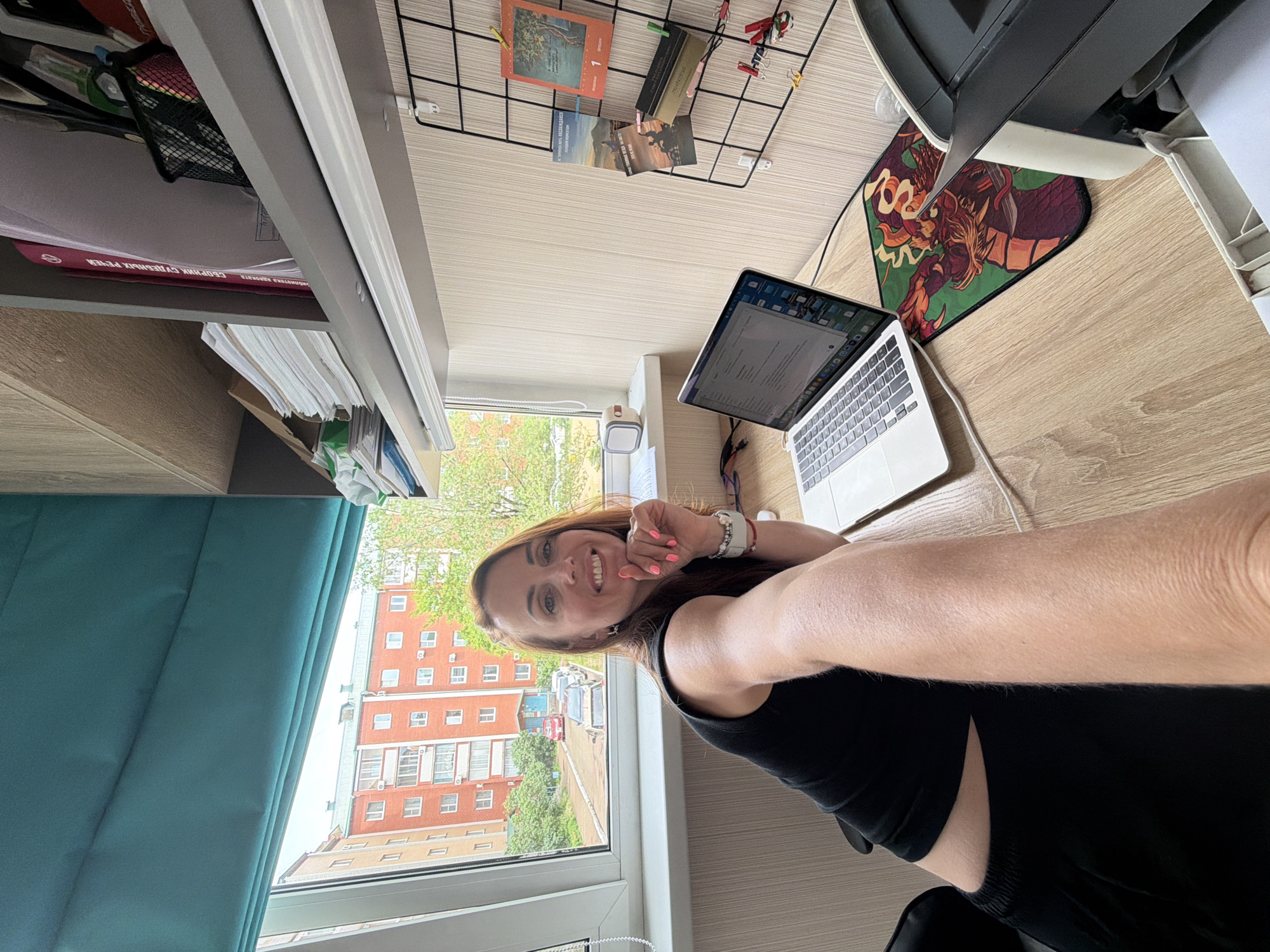 A woman with long auburn hair, wearing a black sleeveless top and black bottoms, is captured in a selfie at a desk. She is smiling and resting her chin on her hand, her nails painted bright pink. A laptop is open in front of her, and a printer is partially visible to the right. The desk is made of light-colored wood. Behind her, a window overlooks a residential area with multi-story brick buildings, trees, and parked cars. A teal-colored roman shade is drawn over the upper part of the window. The background also shows a shelf filled with books and papers, and a black wire grid on the wall adorned with postcards and clips. The scene appears to be an indoor workspace with a view of an urban residential environment in Birobidzhan, Russia.