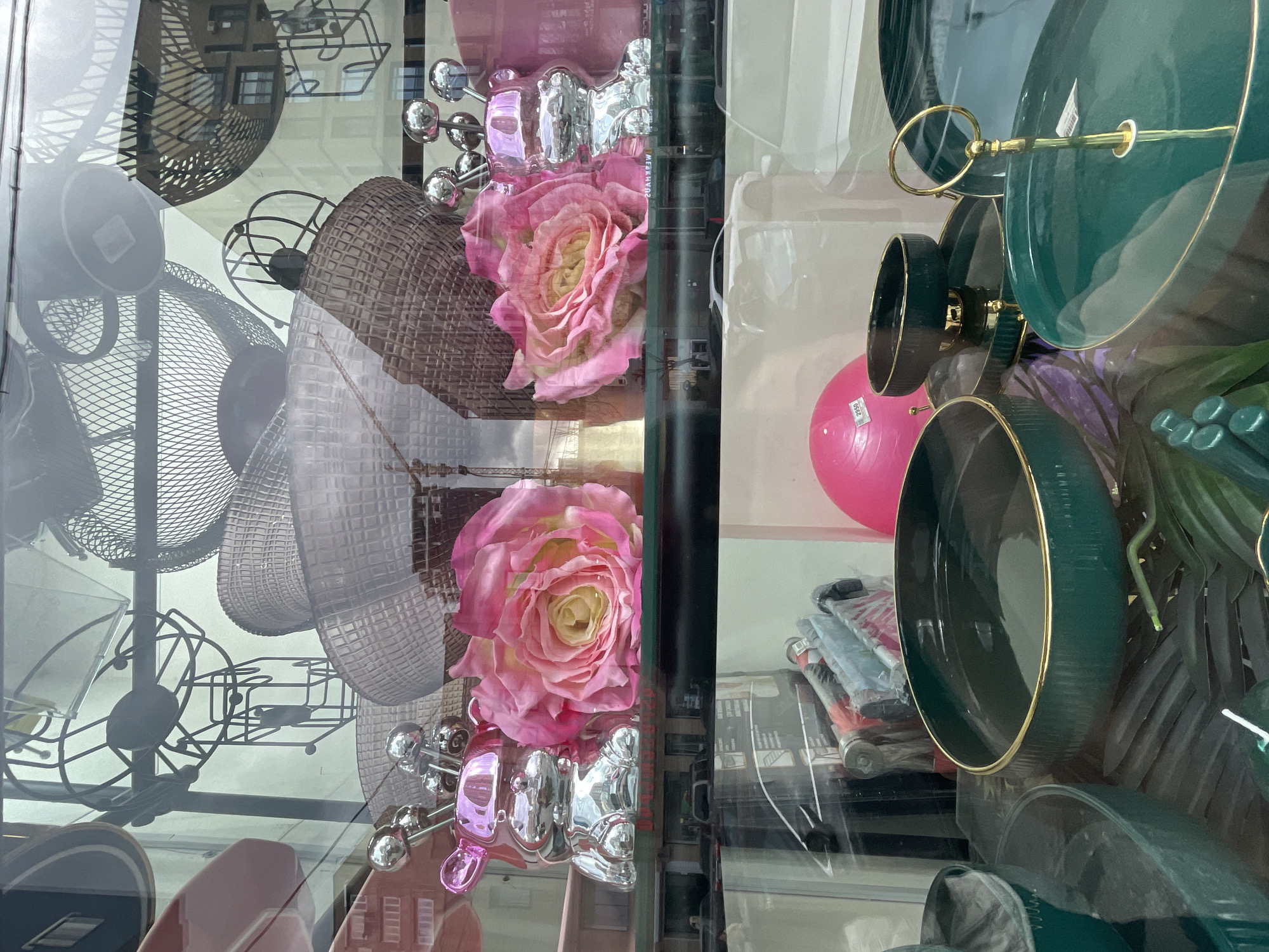 The image appears to be a shop window display in Yerevan, Armenia, as evidenced by the Armenian lettering visible on a sticker. The display features a mix of decorative and household items. On the left side are wire baskets and stands of various sizes and styles. Pink roses are positioned near decorative silver objects resembling bears or dolls. On the right side, there's a stack of green and gold-trimmed bowls and plates. A pink exercise ball is also visible among these items, along with some packaged goods with rollers. The glass of the window is reflecting the building across the street and the city sky.