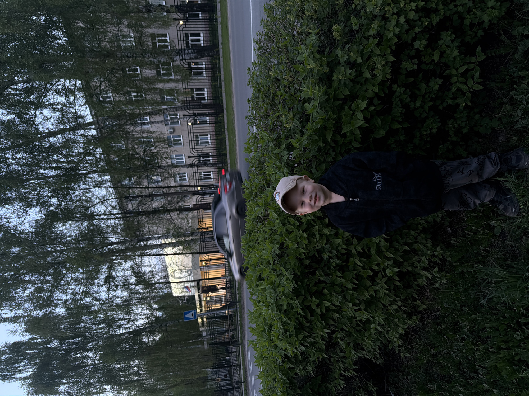 A boy wearing a beige baseball cap, black jacket with "PUMA" branding, and camouflage pants lies in a bush near a street in Chaikovsky, Russia. A silver car with illuminated taillights drives on the adjacent road. On the opposite side of the road, behind a black fence, is a multi-story building with numerous windows. Trees line the street, and a blue and white sign is visible near the road. The environment appears to be dusk or evening.