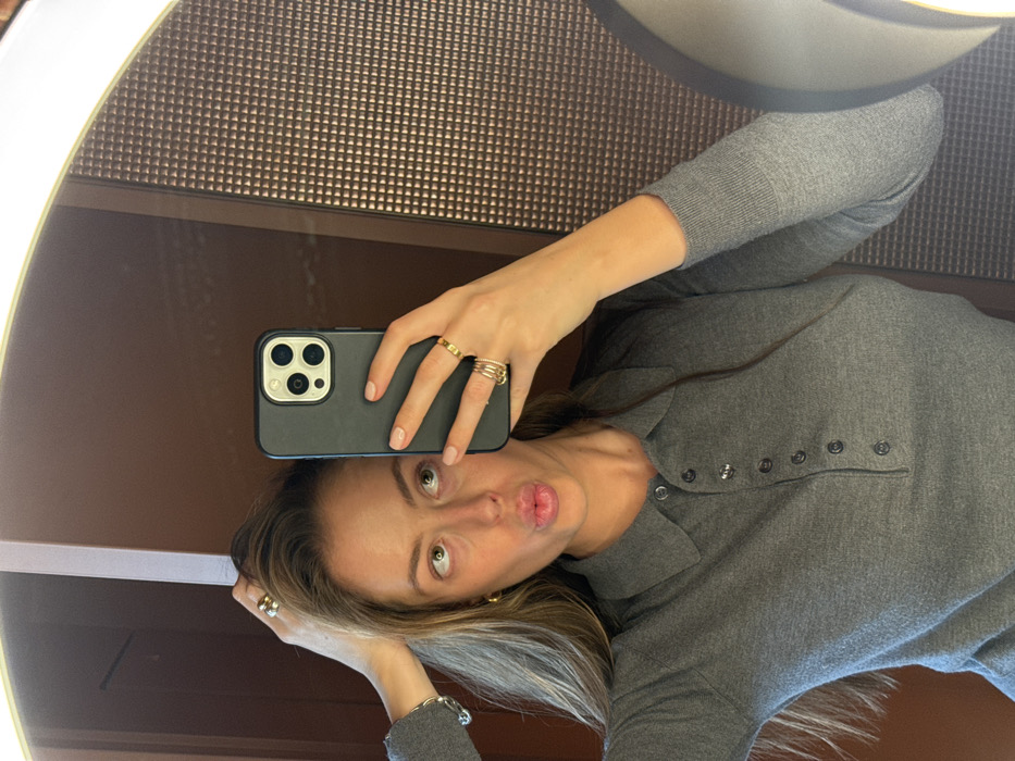 An adult female is depicted in a mirror selfie, posing in an indoor setting. She has long, light brown hair, which she is touching with her left hand, revealing a silver-toned ring on her left ring finger and a silver-toned bracelet on her wrist. Her right hand holds a black smartphone, featuring a triple-lens camera array, positioned to capture her reflection. Her fingernails are painted a light nude color. On her right hand, she wears a gold-toned band ring on her index finger and three stacked gold-toned rings on her ring finger. She wears a dark gray, long-sleeved polo shirt with a buttoned placket. Her facial expression shows pursed lips and her eyes are looking upwards and slightly to the left. The background reflected in the mirror behind her head consists of a plain brown surface on the left and a dark brown wall panel with a textured, repeating square/pyramid pattern on the right. An illuminated light strip is visible along the top edge of the circular or oval mirror frame. The location is Unknown, Unknown.