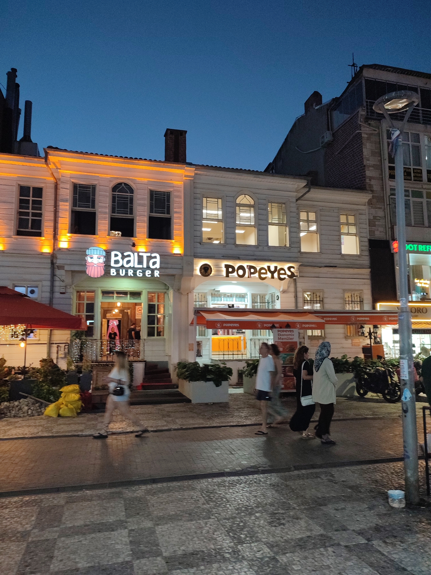 The image depicts a street scene in Kadıköy, Turkey, at dusk or early night. The main focus is on the facades of two adjacent establishments, a "BALTA BURGER" restaurant and a "POPEYES" fast-food chain. Both buildings have white exteriors with orange accent lighting. BALTA BURGER features a cartoonish logo with a bearded character. Popeyes has a prominent orange sign and an orange awning.
Several people are walking on the cobblestone sidewalk in front of the establishments. One woman, blurred due to motion, is walking towards the left, wearing a white top and dark shorts. To her right, a group of three people, two women and a man, are walking towards the right. One woman is wearing a headscarf and a long beige coat over dark pants, carrying a white purse. The other woman is wearing a black dress. The man is wearing a white t-shirt and dark shorts.
To the left of BALTA BURGER, there is a red umbrella covering an outdoor seating area, with some string lights visible. Yellow bags are piled near the entrance. To the right of Popeyes, a black motorcycle is parked. A streetlamp with a modern design stands near the Popeyes establishment, and a traffic light is visible further down the street. The sky above is a deep blue, indicating the transition from day to night. The overall atmosphere is that of a busy urban commercial area.