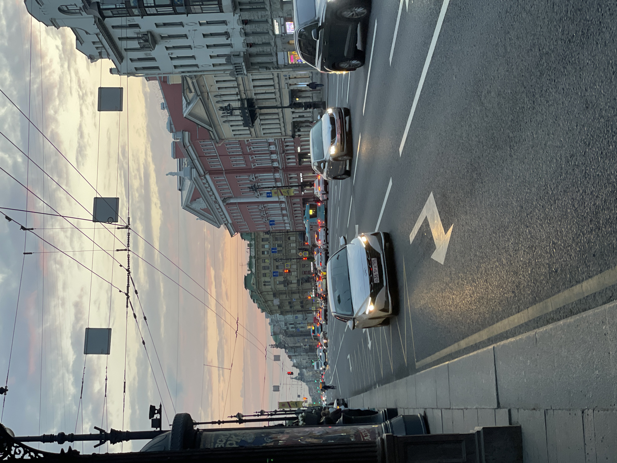 The image shows a street in Saint Petersburg, Russia, at dusk. The street is a multi-lane road with cars driving in both directions. A white arrow pointing left is painted on the road. Buildings line both sides of the street, with architectural styles typical of Saint Petersburg. Overhead, a complex network of wires and electrical fixtures spans the street. The sky displays colors of sunset, with pale oranges and pinks blending into gray clouds. Sidewalks are visible on the sides of the street, with a low stone barrier along the edge. Cars are the main objects in motion, with their headlights illuminated. A few distant pedestrians are visible. The overall atmosphere is urban and somewhat congested.