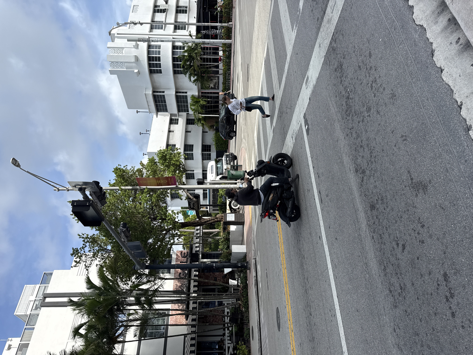 The image captures a street scene in Miami Beach, Florida, United States. The street runs from the lower left to the upper right of the image. A person on a black scooter is on the right side of the street. A woman is crossing the street, walking leftward, with a black car directly behind her. Several white multi-story buildings line the left side of the street, interspersed with palm trees. Traffic lights and street signs are also visible on the left side. The road markings include white lines and yellow lines.