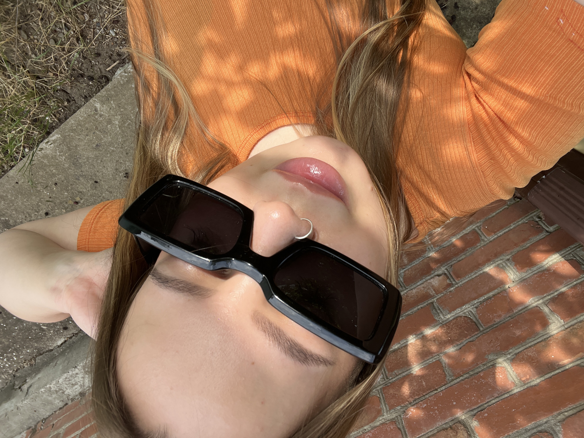 An eye-level, close-up, selfie-style, outdoor shot shows a young Caucasian woman wearing oversized black rectangular sunglasses and an orange ribbed t-shirt. Her hair is blonde and appears to be shoulder-length. She is wearing a silver hoop nose ring. The background is blurred, showing a brick wall on the left and a concrete surface with some dry grass and dirt on the right. The lighting suggests it is daytime, with dappled sunlight creating shadows on the brick wall. The image does not provide any specific clues about the location in Novopokrovskaya, Russia, or any discernable urban elements.