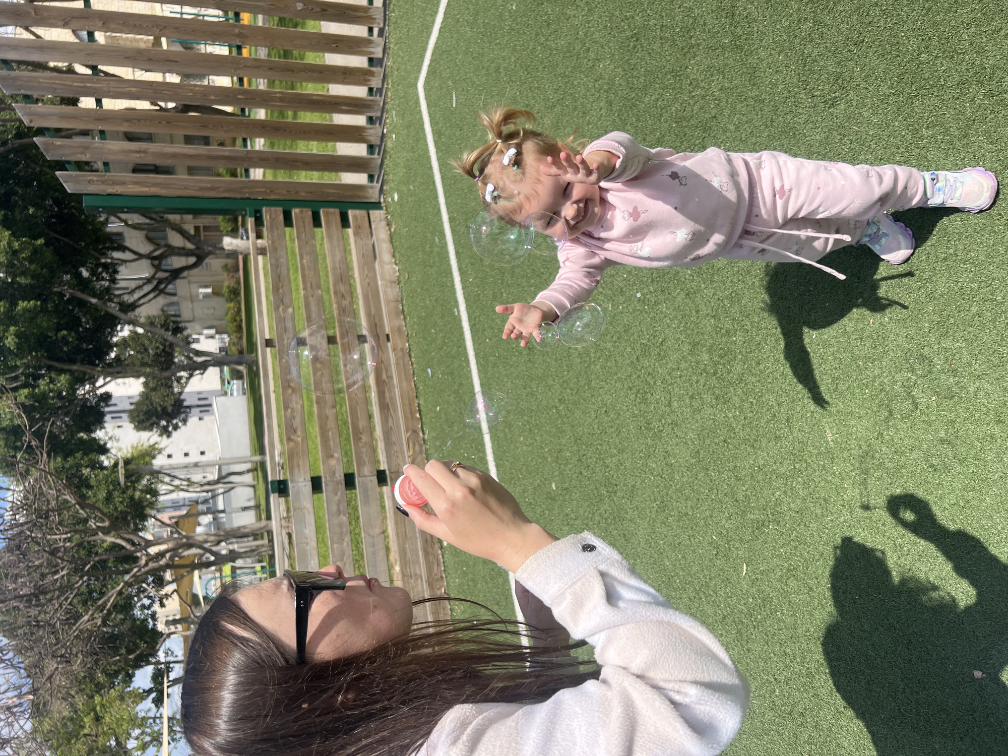 A woman blows bubbles for a toddler on a green synthetic surface, possibly a playground. The woman, wearing a light-colored long-sleeved shirt and dark sunglasses, is holding a pink bubble wand. The toddler, in a pink tracksuit with ballerina figures, reaches for the bubbles with a smile. The background features a wooden fence, trees, and a white building. The scene is outdoors, likely in Kiryat Bialik, Israel. The image is taken from a high angle, looking down at the subjects. Shadows are visible on the green surface.