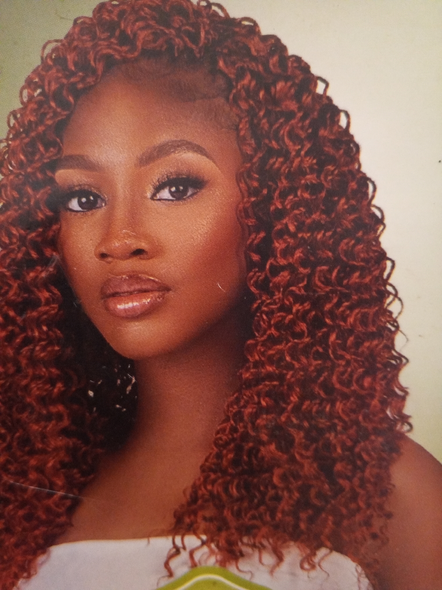 A close-up portrait of a woman with dark skin and curly, red-brown hair. She has dark eyes, well-defined eyebrows, and glossy lips. She is wearing makeup, including eyeliner and eyeshadow. She is wearing a white garment with green stitching near the bottom. The background is a blurry, neutral color. The location in Gwong, Nigeria is unknown.