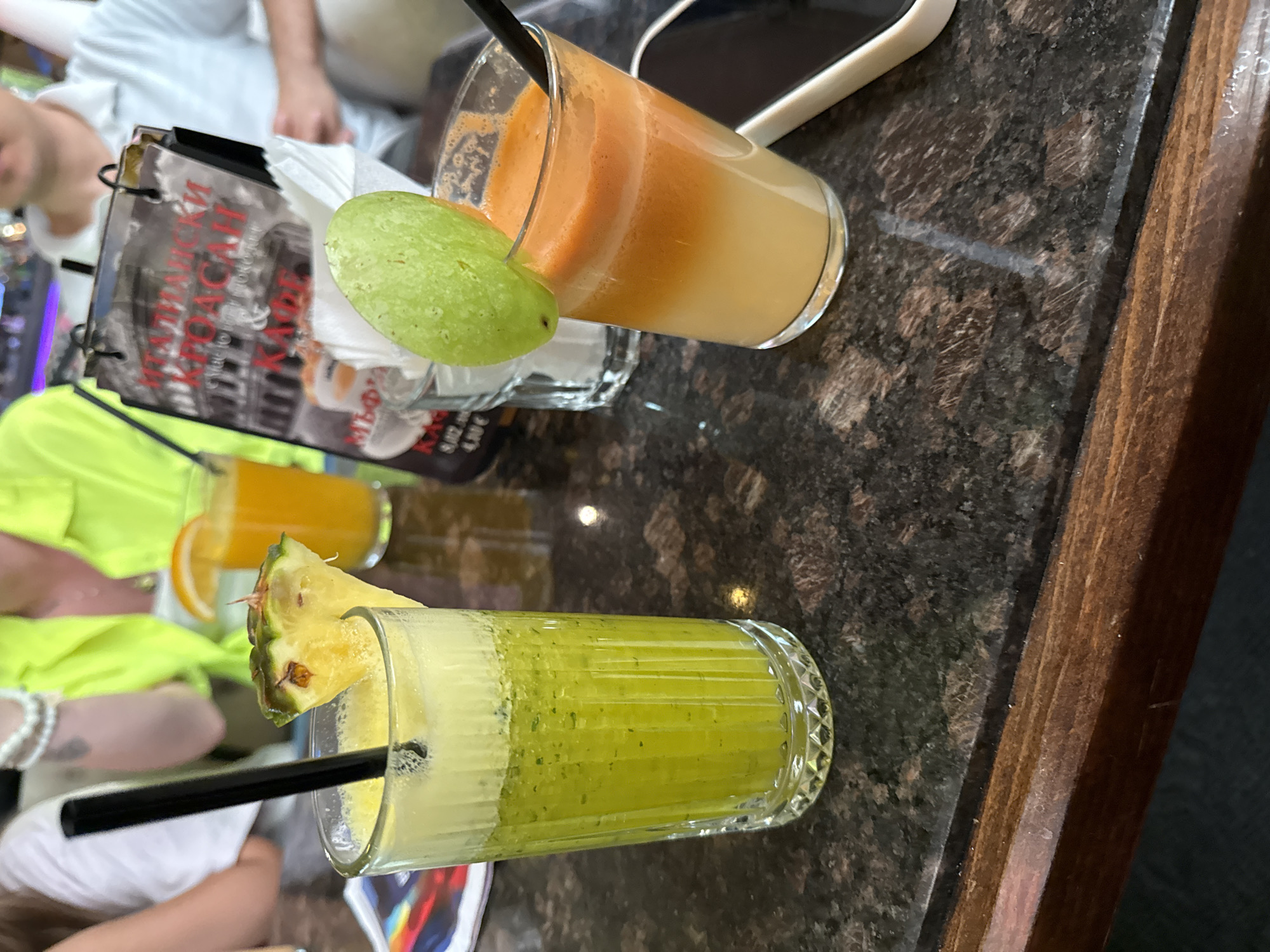 On a dark marble tabletop with a wooden edge, two glasses containing beverages are visible. The glass at the top contains a layered orange and yellow drink, garnished with a green fruit slice, and has a black straw. A black cell phone is positioned behind it. The second glass, at the bottom, holds a green drink topped with a pineapple wedge and a black straw. Both glasses are partially reflected in the tabletop. In the background, there are people and items that suggest a cafe or restaurant setting. The location appears to be a public space with people dining. The city and country are not discernible from the image.