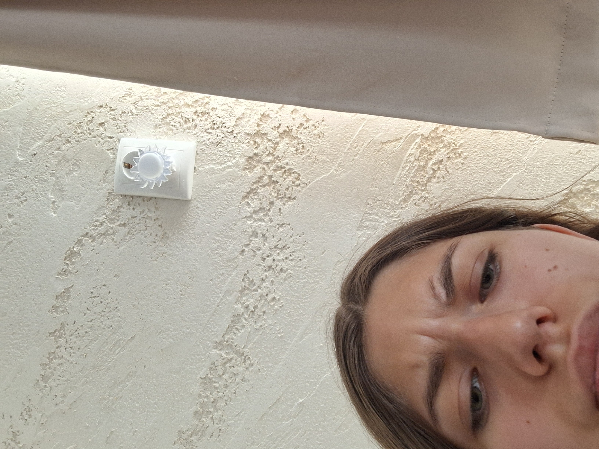 A low-angle, close-up shot shows the upper portion of a young woman's face and the wall behind her, which is textured and painted a light cream color. On the wall, a white electrical device with a sun-shaped decorative cover is mounted to the right of the woman's face. To the right of the electrical device, a light beige curtain hangs vertically, obscuring a portion of the background. The woman's eyebrows are furrowed, and her eyes are looking downwards, suggesting an expression of confusion or concern. The image appears to be taken indoors, likely in a residential setting in Panagyurishte, Bulgaria, as indicated by the specific metadata.