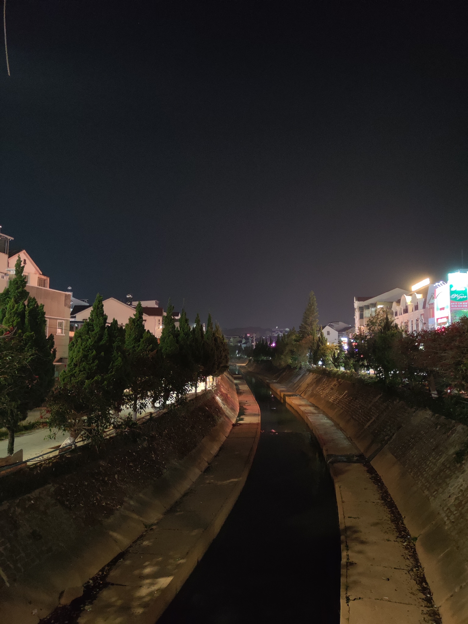 The image depicts a concrete canal in Da Lat, Vietnam, at night. The canal is narrow and appears to contain a small amount of dark water. The concrete sides of the canal are visible, showing signs of wear and fallen leaves. Along the canal banks, there are trees and buildings. Some buildings are illuminated, contributing to the city's night glow. The sky is dark, with little visible detail. No people are visible in the scene.