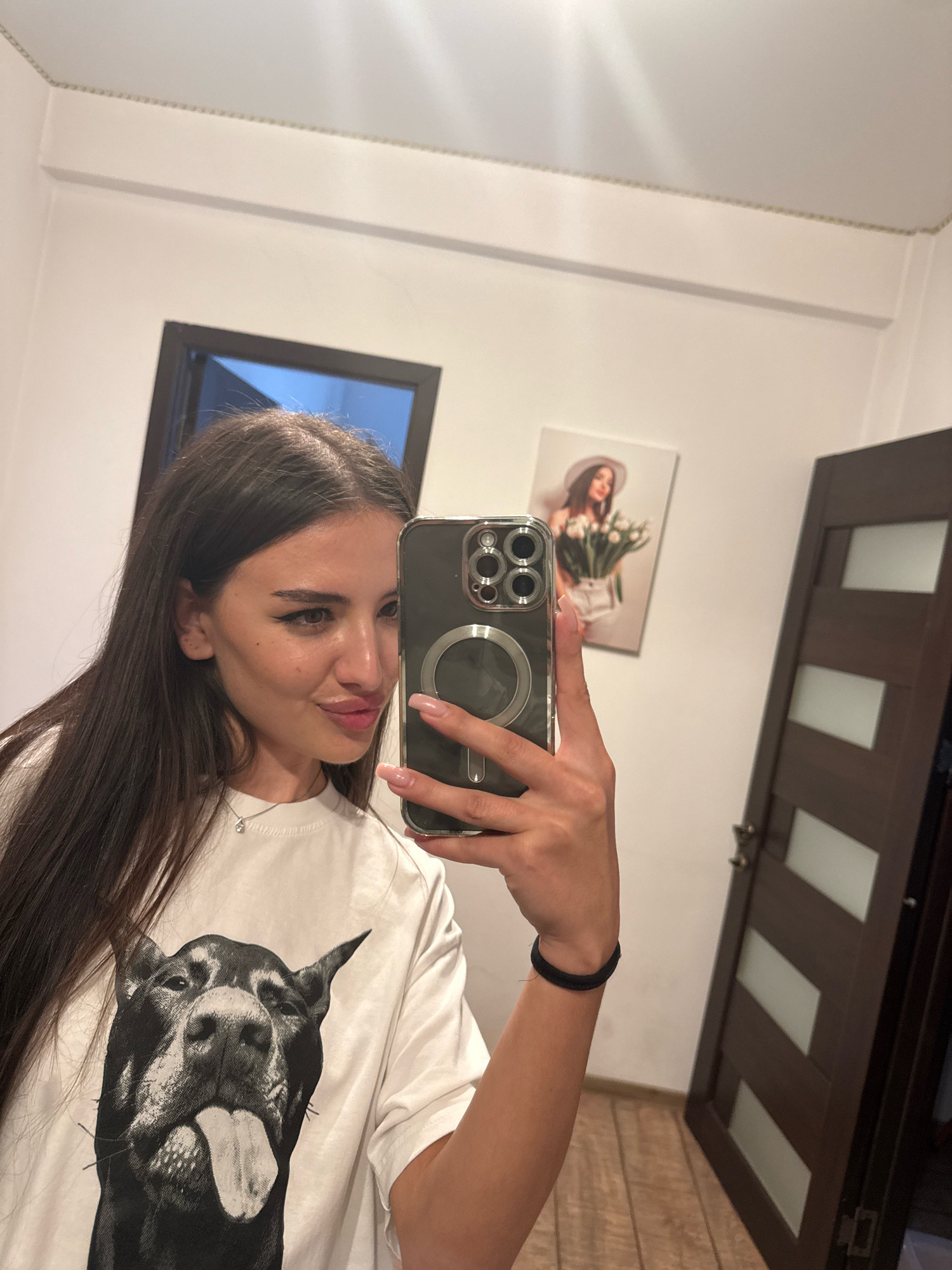 A young woman with long, dark hair is taking a selfie in a room. She is wearing a white t-shirt with a large black and white print of a dog's face. She has on a thin silver necklace with a small pendant. Her left hand is holding a smartphone in front of her. The background shows a white wall, a framed picture of a woman holding flowers, a dark wooden door, and a glimpse of a doorway. The floor appears to be made of wood.