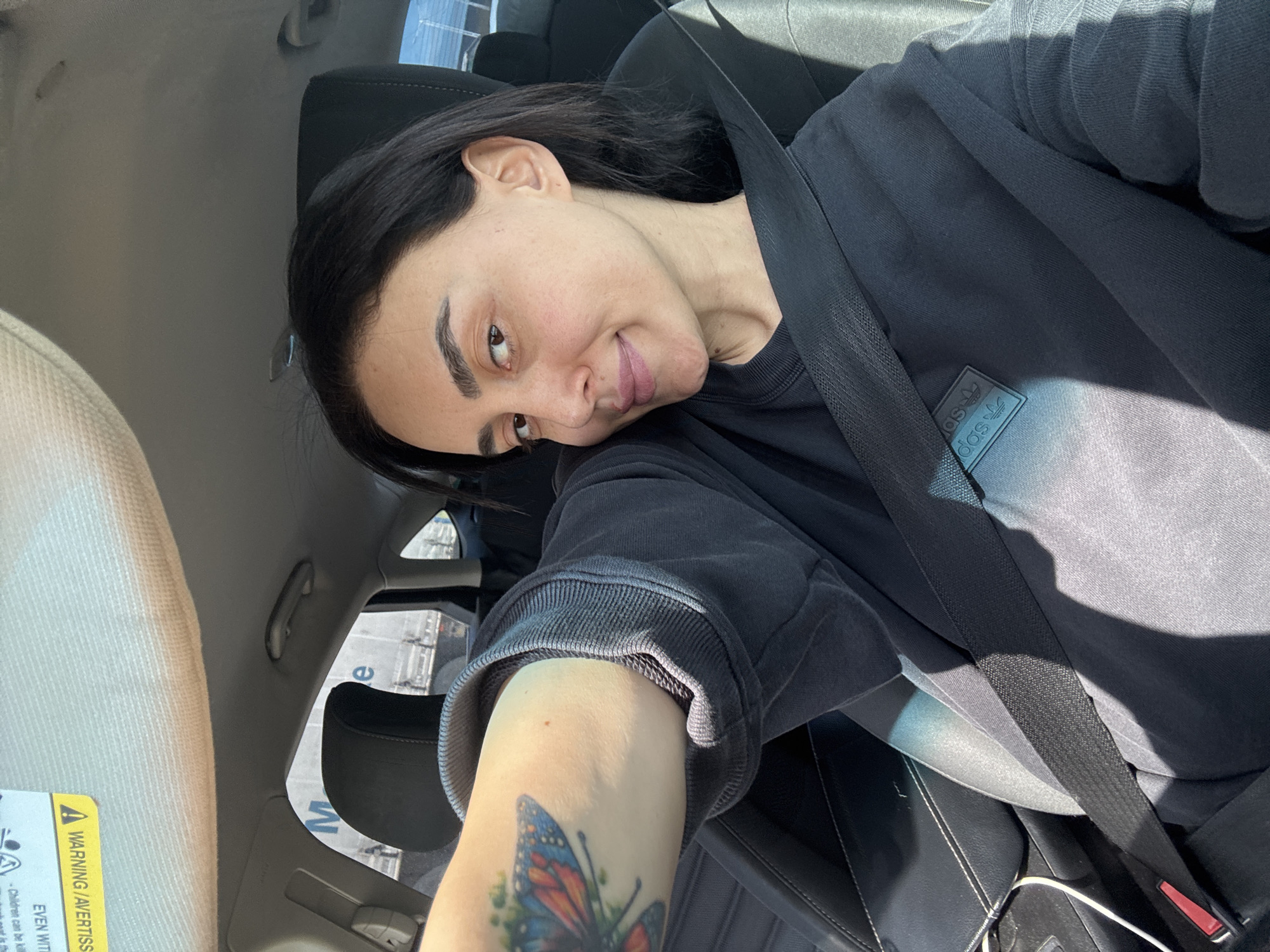 A woman is captured in a selfie-style shot from inside a car, likely in Tbilisi, Georgia. She is positioned in the driver's or passenger's seat, looking towards the camera with a slight smile or smirk and her head tilted. She has dark hair, dark eyebrows, and a fair complexion. She is wearing a dark grey or black long-sleeved sweatshirt, featuring a faint "adidas" logo on the chest. A seatbelt diagonally crosses her torso.
Her left arm is visible, extended towards the camera, revealing a colorful butterfly tattoo on her forearm. Bright sunlight streams into the car, creating highlights on her skin and clothing, and casting shadows within the interior.
The car's interior shows a grey headliner, a light-colored sun visor on the left with visible warning labels stating "WARNING / AVERTISSEMENT", and dark car seats. Through the rear window, a building or structure is visible with partial text, including the letters "e" and "M". The overall impression is a casual, daytime moment.
