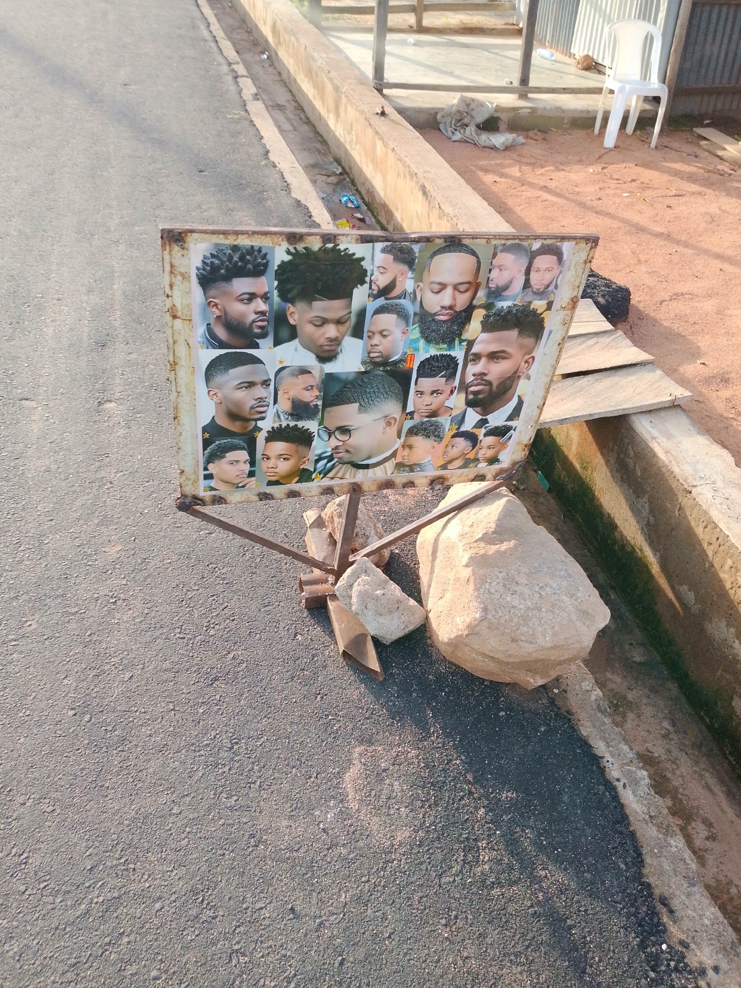 The image depicts a weathered, rectangular sign displaying various hairstyles for men, likely from a barber shop. The sign is propped up by a rusty metal stand, which is partially secured by two large rocks placed on either side of its base. The sign is situated outdoors, on the edge of a paved surface that appears to be a roadside or walkway, bordering a dirt area and a concrete drainage channel. In the background, there is a white plastic chair and some debris, suggesting an informal commercial or residential area. The lighting indicates it is daytime, with sunlight casting shadows. The overall scene appears to be in Jalingo, Nigeria, given the context.