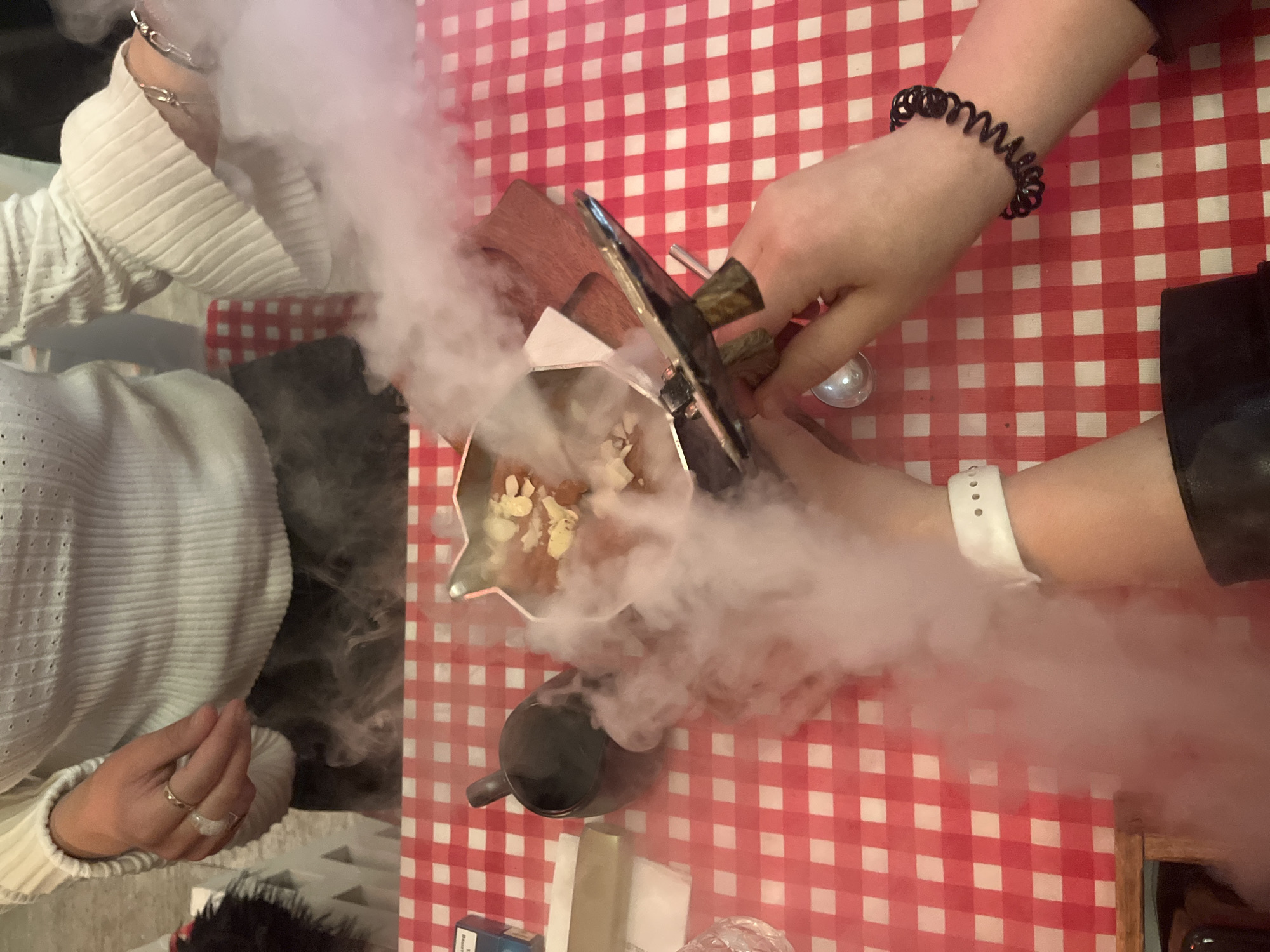 An overhead shot captures a dining scene on a table covered with a red and white checkered tablecloth. Two individuals are present, primarily represented by their hands and forearms.
On the left, a person wears a white ribbed long-sleeve top, a silver chain-link bracelet on the wrist, and rings on their fingers. On the right, another person wears a black top or sleeve, with a black coiled hair tie and a white smartwatch visible on their wrist.
The central focus is a theatrical food presentation, likely a dessert. A metal serving dish or bowl contains a dish topped with nuts or crumbs, surrounded by a dense cloud of white smoke or vapor, likely from dry ice. The person on the right is actively using metal tongs to interact with the smoky dish.
Other objects on the table include a small black mug, a wooden serving board, a reflective spoon or small dish, and a blue and white packet, faintly showing "Marlboro" text, along with other small, indistinct items.
The scene suggests an indoor setting, possibly a restaurant or cafe in Plovdiv, Bulgaria, where a visually engaging dessert or cold dish is being served. The time of day is not discernible, and no specific emotions are visible, but the presentation implies an enjoyable and novel dining experience.