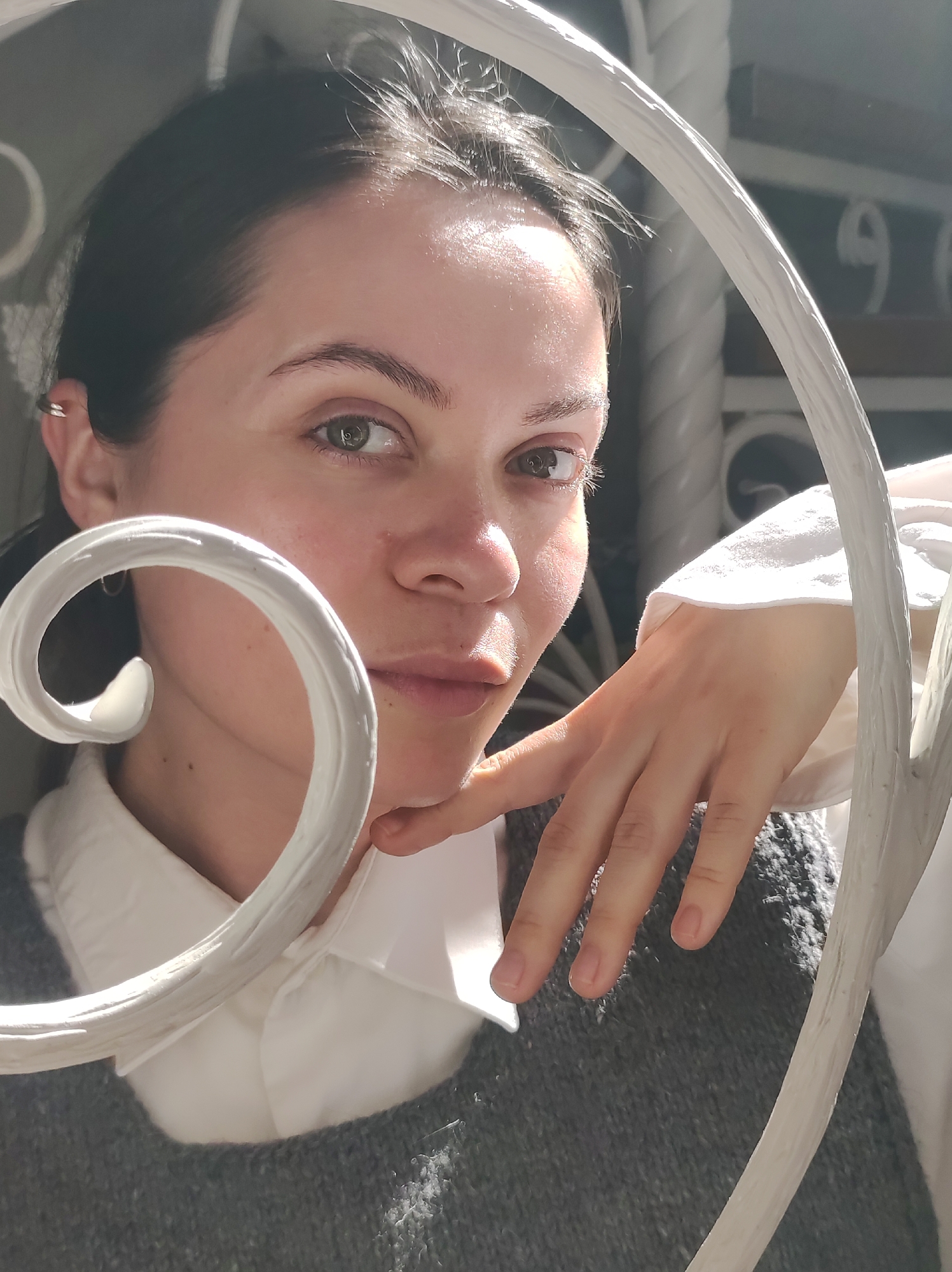 A young woman with dark hair, fair skin, and hazel eyes looks directly at the camera. She is wearing a white collared shirt underneath a grey knit sweater. Her right hand is resting on her chin. The background features a white wrought-iron railing. Through the railing, numbers, possibly building numbers, are visible, partially obscured by the railing structure. Based on the prompt's instruction, this scene is set in the city of Фоки, Russia.