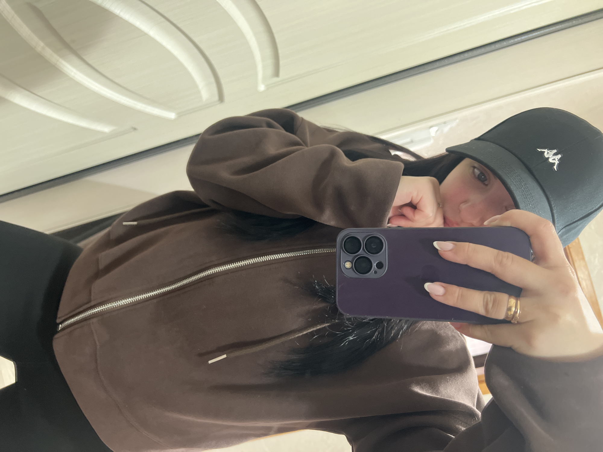 A woman takes a selfie in what appears to be an indoor setting. She is wearing a dark baseball cap with a Kappa logo, a brown zip-up hoodie, and what looks like black leggings. She holds a purple iPhone in her right hand, obscuring part of her face. Her hair is long and dark. Her left hand rests near her face. The background features a white door with curved paneling. The photograph seems to be taken in Марковский, Russia.