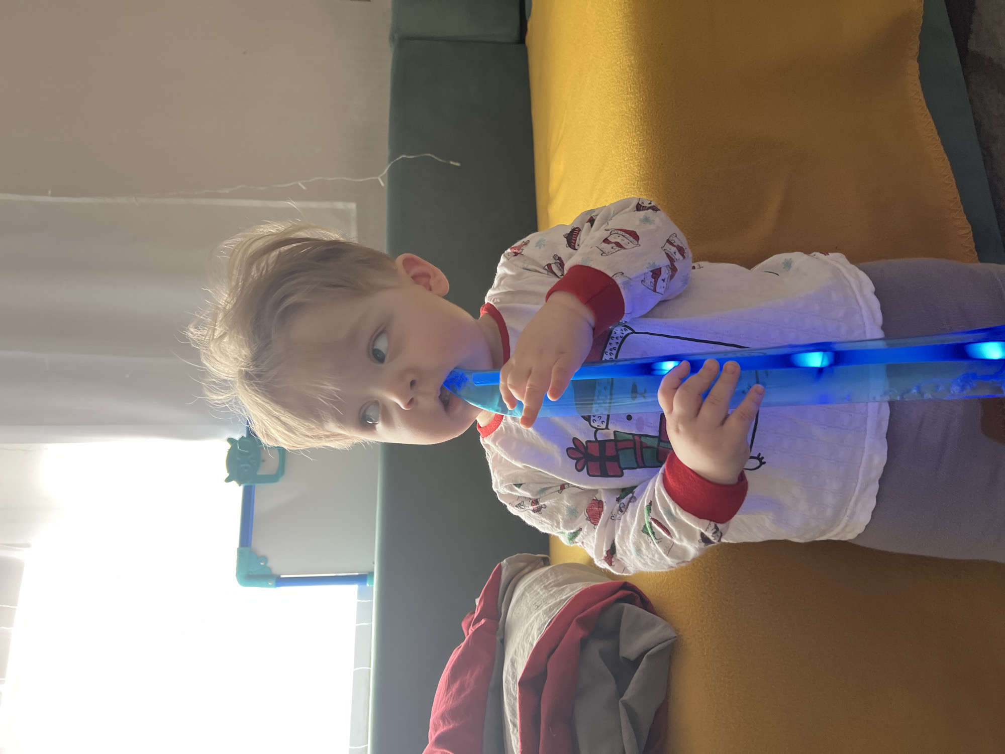 A candid, vertically oriented shot captures a young child indoors. The child, with light blonde hair, is wearing white pajamas adorned with festive patterns of snowmen and wrapped presents, featuring red cuffs. They are holding a transparent blue, light-up toy that resembles a flute or a long tube, with one end near their mouth, suggesting they are either playing it or teething on it. The child looks towards the camera with an engaging and slightly curious expression.
The background includes a couch with dark green and yellow cushions. A pile of red and grey fabric is visible on the yellow cushion. To the left, a bright light source, likely a window, illuminates the room, and part of a white board with a green frame is visible. The scene is brightly lit, indicating daytime. There is no discernible text in the image.