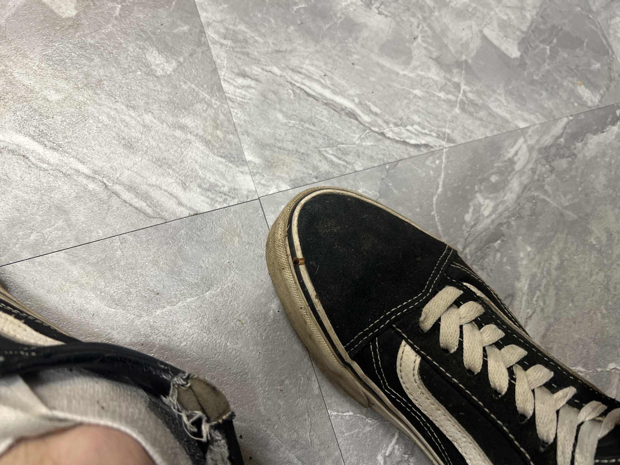 The image shows a close-up, overhead view of two worn, black Vans sneakers on a gray, marbled tile floor. One shoe, on the left, is partially visible and appears to have a torn section near the ankle opening, revealing white fabric underneath. The other shoe, more prominently featured on the right, shows the toe box and laces of a black canvas Vans sneaker with a white side stripe and white laces. The white rubber sole of this shoe is dirty and shows signs of wear. A thin, dark line, likely a grout line between tiles, runs diagonally across the image. There are no people visible, and the scene appears to be indoors. The image provides no information about the location in the city of Ленинский, Russia.