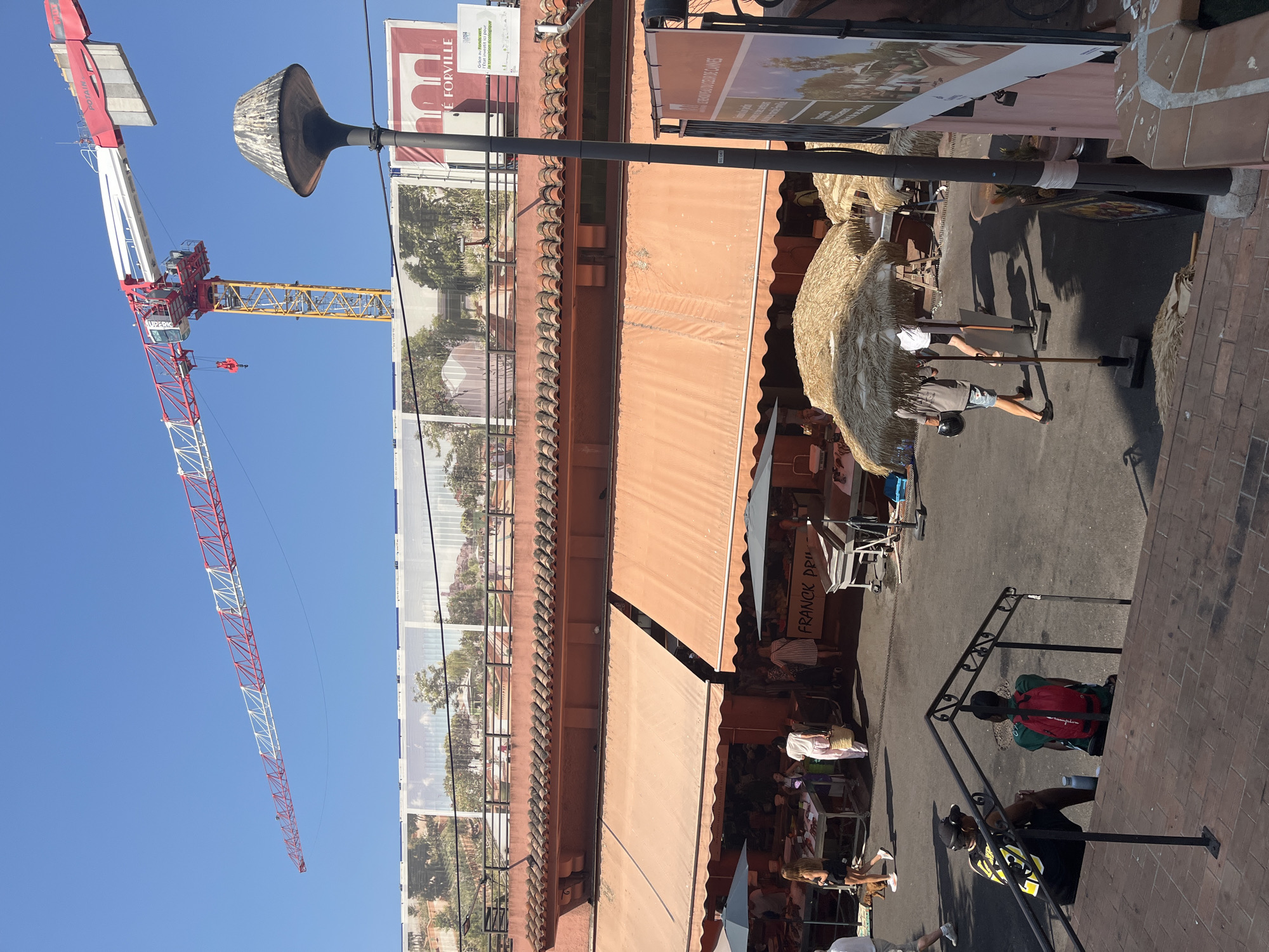 A wide-angle, eye-level shot captures a street scene in Cannes, France, dominated by a construction crane and a market area. The sky is a clear, bright blue. The crane, a large red and white Potain model, extends diagonally across the upper portion of the frame. Below the crane, a large billboard or banner displays architectural renderings of buildings and landscaping.
In the mid-ground, the facade of a market building is visible, featuring orange awnings and terracotta roof tiles. Several stalls are set up beneath the awnings, with people browsing and interacting. One stall prominently displays the name "FRANCK PRI". Straw-thatched umbrellas provide shade for some of the market stalls. Various individuals are present in the market area, some standing, some walking.
In the foreground, two young men are seated on steps, facing away from the camera. One wears a black and yellow shirt and the other a green shirt with a red backpack. A black metal railing runs along the steps. The ground is paved with bricks and asphalt. A lamppost stands to the right of the frame. The overall impression is of a bustling urban environment with ongoing development.