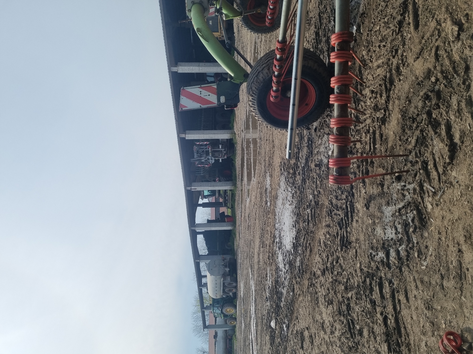 The image shows a Claas Volto tedder parked on a muddy dirt surface in Ovcharovo, Bulgaria. Behind it, there is a shed with farming machinery inside. A light-gray sky is visible above.