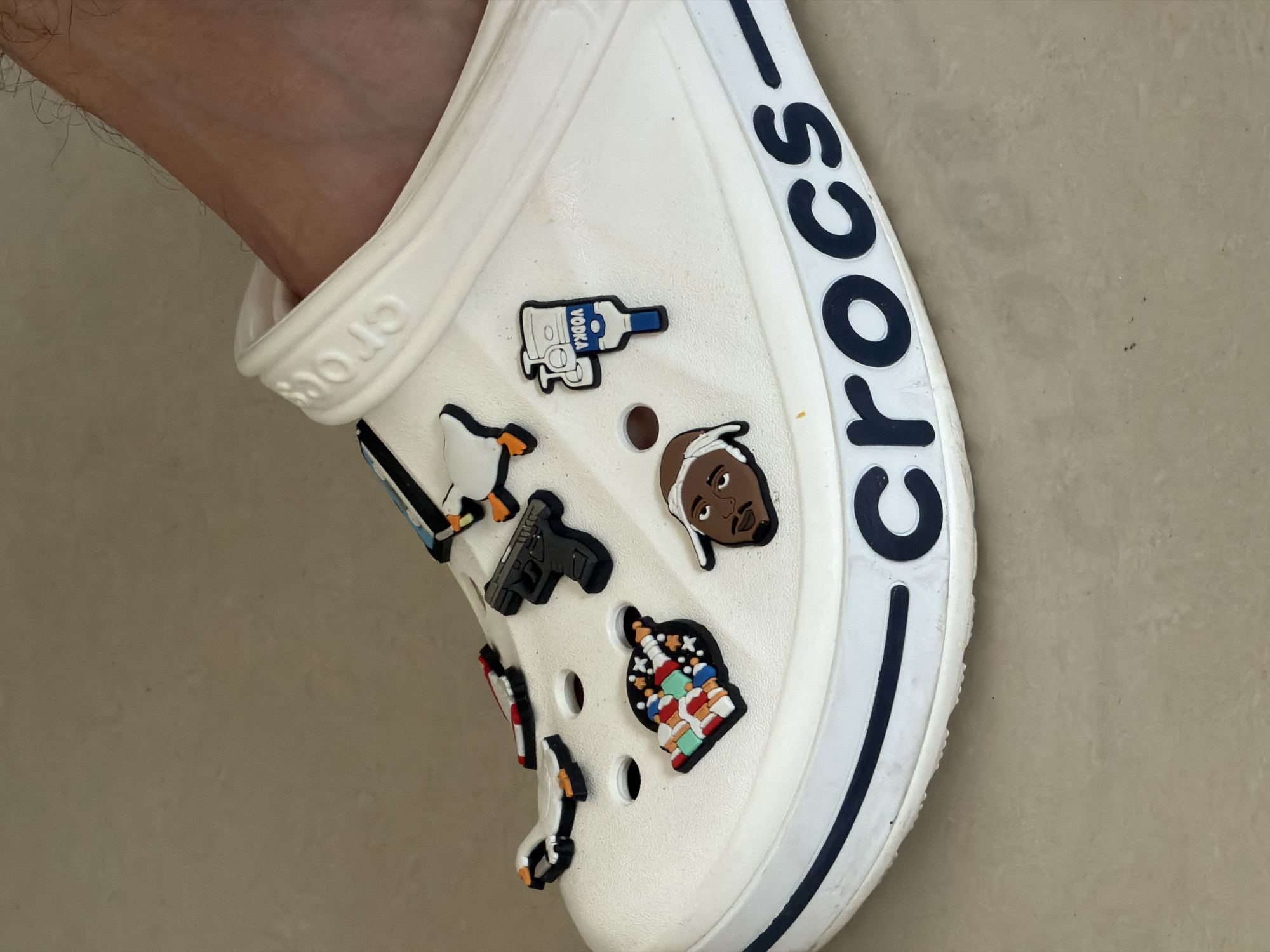 A close-up shot shows a white Croc shoe decorated with various charms. The charms include a bottle with "VODKA" written on it, a penguin, a gun, a depiction of a Black man, and a colorful building. The word "Crocs" is displayed in large, blue letters on the side of the shoe. A person's leg is visible near the shoe. The background is a light-colored, neutral surface.
