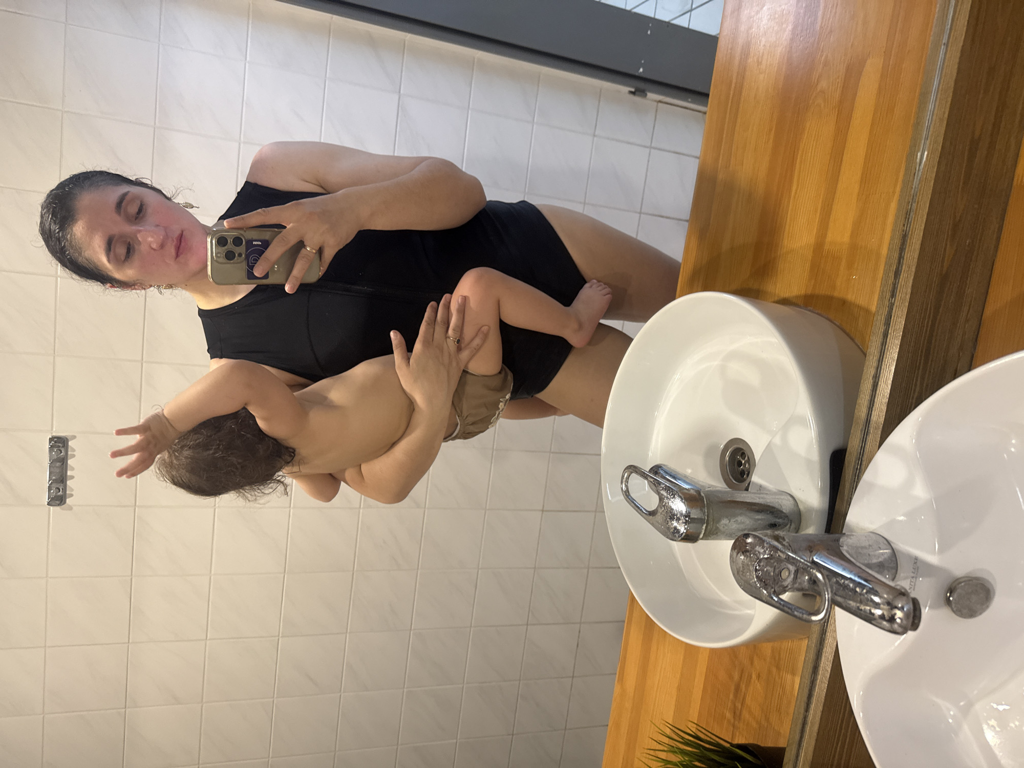 A woman is holding a baby in a bathroom and taking a selfie in the mirror. The woman is wearing a black one-piece swimsuit. The baby is facing away from the camera and wearing a diaper. The bathroom has white tiled walls and a wooden countertop with two white sinks. The location is possibly Tobolsk, Russia, based on the sinks' brand name "AITA LUXE" visible in the mirror.