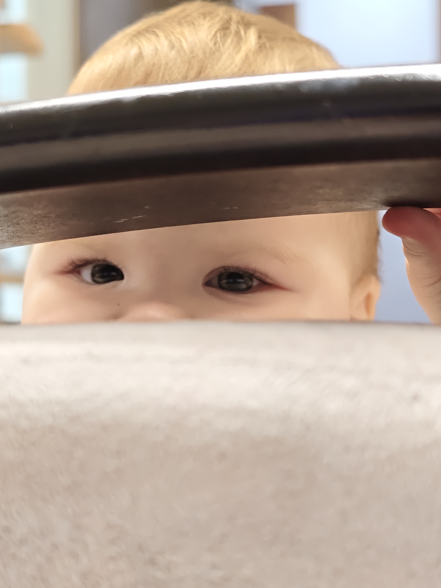 The image captures a close-up, low-angle perspective of a baby or toddler peeking over a dark, horizontal rail or bar, in Chaikovsky, Russia. The primary subject is the baby, whose blonde hair and alert, dark eyes are visible above the rail. The rest of the baby's face is obscured by the bar. A small hand, likely belonging to the baby, is seen on the right side, gripping the top of the bar.
The activity depicted is the baby curiously looking over an obstacle, perhaps a crib rail, playpen, or a piece of furniture. The wide-open eyes suggest an inquisitive and observant demeanor.
In the foreground, a blurry, lighter-colored, textured surface extends across the bottom of the frame, further suggesting the baby is positioned behind a barrier. The background behind the baby is soft and out of focus, showing indistinct light areas and a hint of light blue on the right, indicating an indoor setting, likely a home or nursery, under soft daytime lighting. There is no visible text in the image.