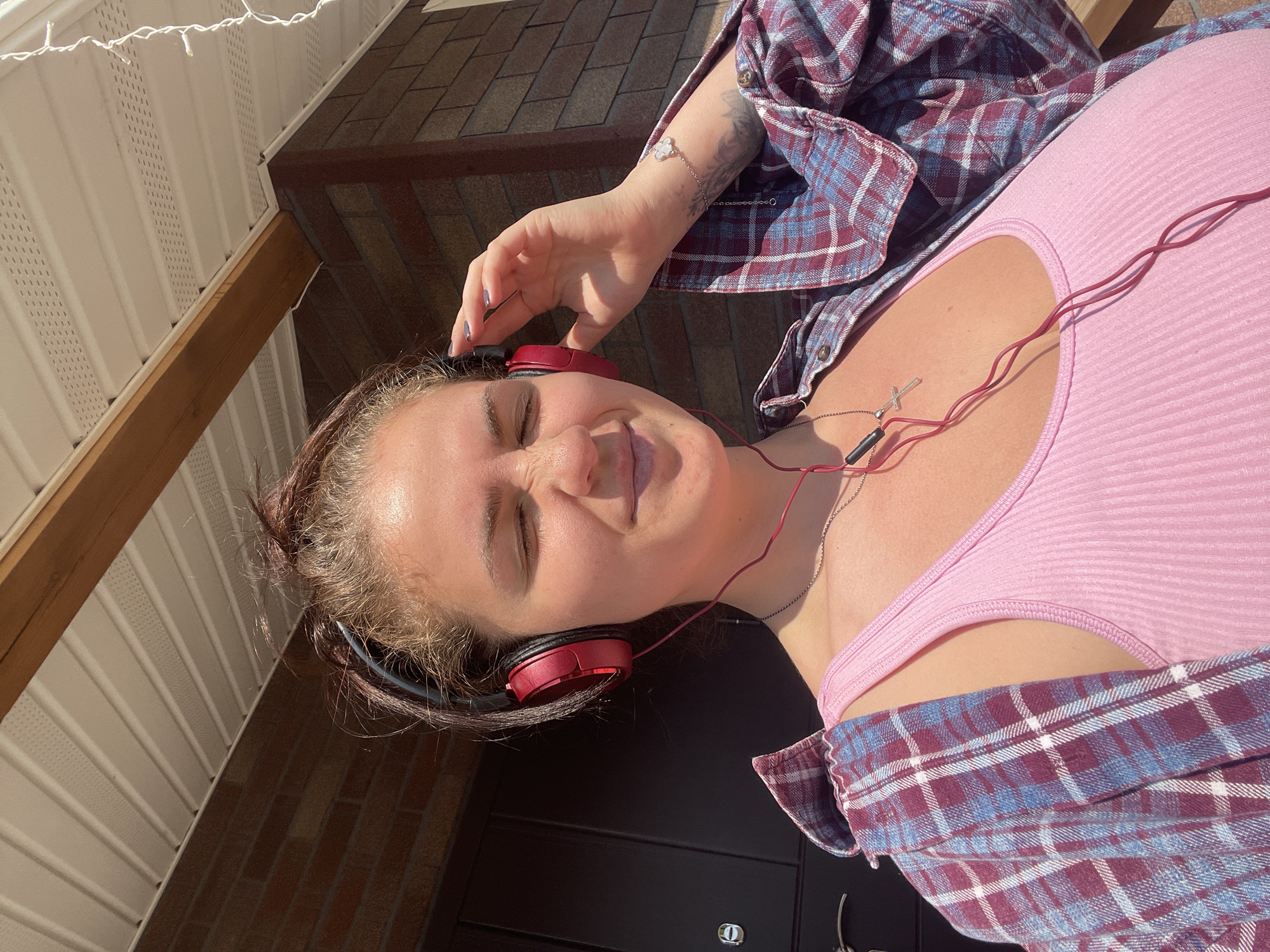 The image, taken in Мосметаллоконструкция Онт, Russia, is oriented sideways, showing a young woman lying horizontally outdoors on a sunny day. She wears a pink ribbed tank top and a maroon and blue plaid shirt unbuttoned over it. Red over-ear headphones rest on her head, and red wired earphones with a black control component are draped around her neck. A silver necklace with a small cross pendant is visible, along with a silver bracelet adorned with a white, possibly clover-shaped charm on her left wrist, which also shows a partial tattoo. Her eyes are closed, and a subtle smile plays on her lips, suggesting a relaxed and content mood. Her right hand is raised near her head, perhaps adjusting the headphones or resting.
The background features the exterior of a building, likely a residence. Above the woman, white vinyl soffit is visible, with white string lights strung along it. A wooden beam runs diagonally across the frame, leading up to a roof covered in brown shingles. Below her, part of a dark brick wall and a dark door with a silver keyhole are visible. The bright sunlight creates strong highlights and shadows, indicating a clear, sunny daytime setting, possibly late morning or afternoon. There is no visible text in the image.